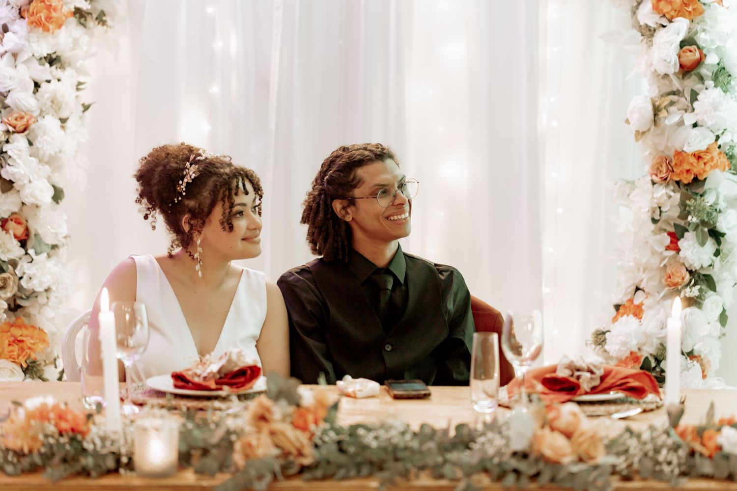 Wedding couple sitting at elegantly decorated reception table with peach floral arrangements and white drapery.