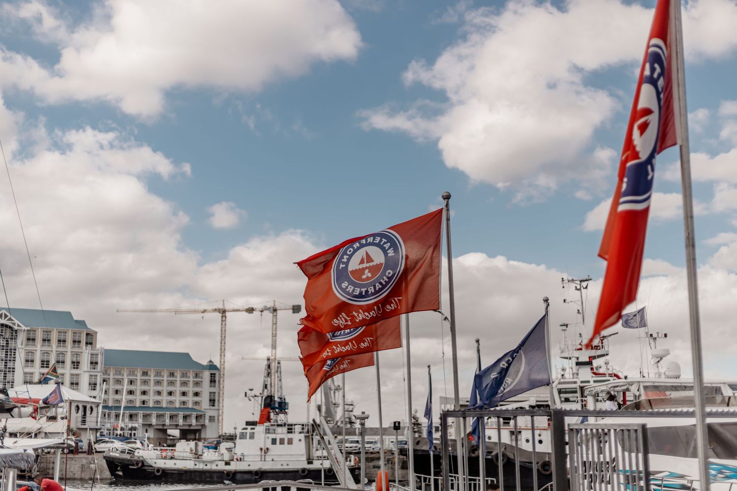 Red flags wave in the breeze at a marina with boats and buildings visible along the waterfront on a cloudy day.