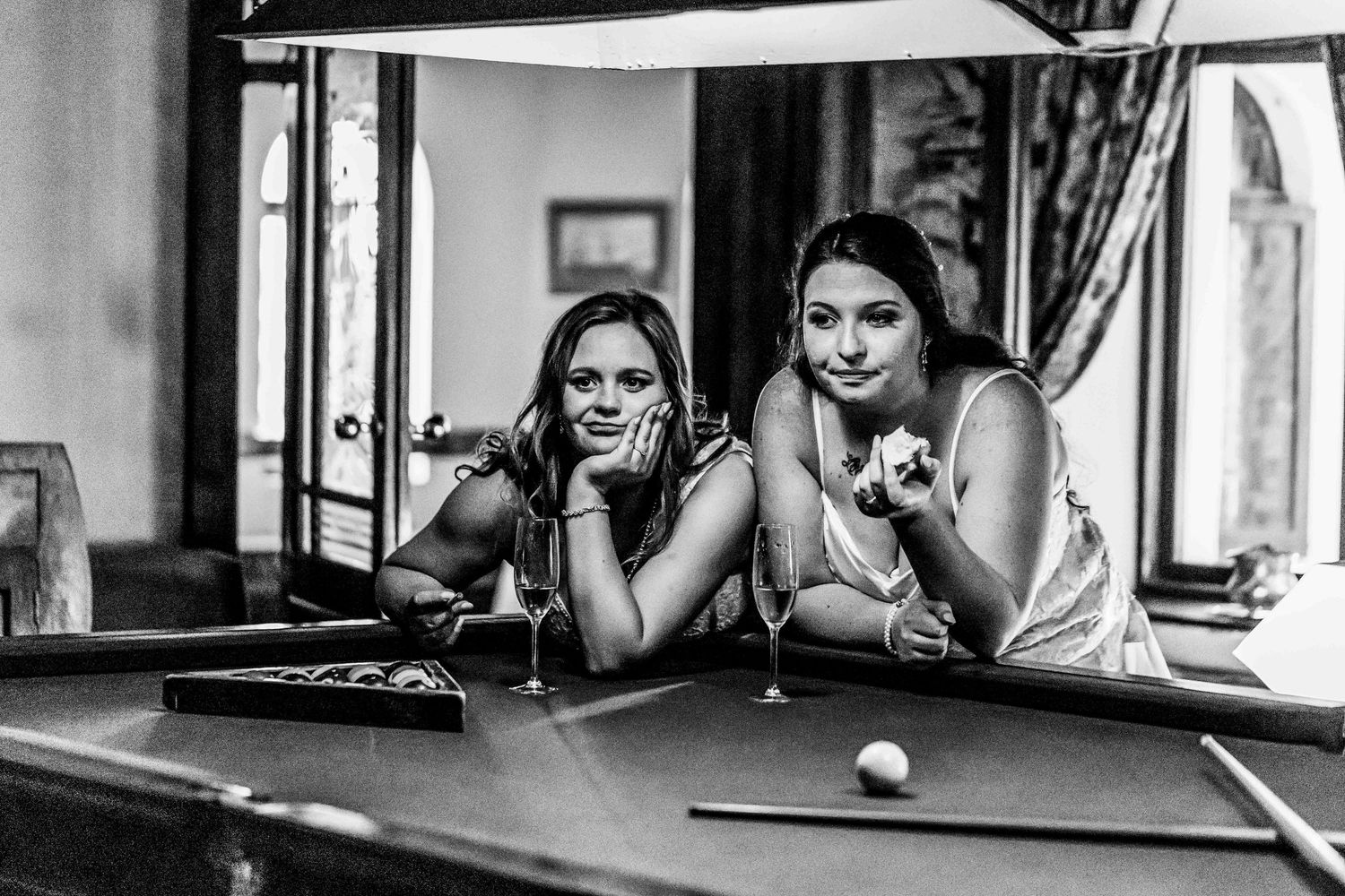 Two women playing pool and laughing together at a billiards table in a black and white monochrome photograph.