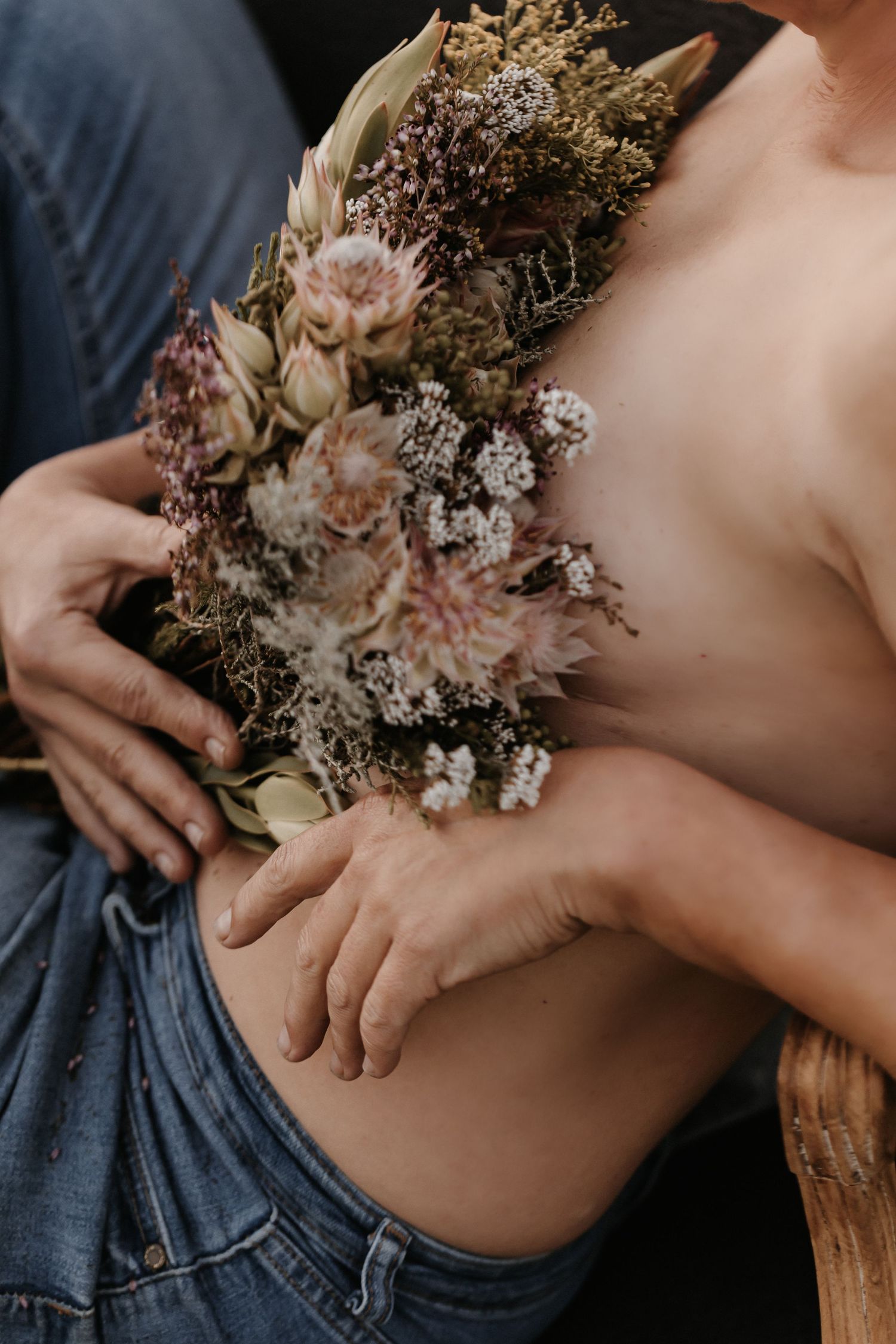 A delicate dried flower bouquet held against bare skin and denim in moody natural lighting.