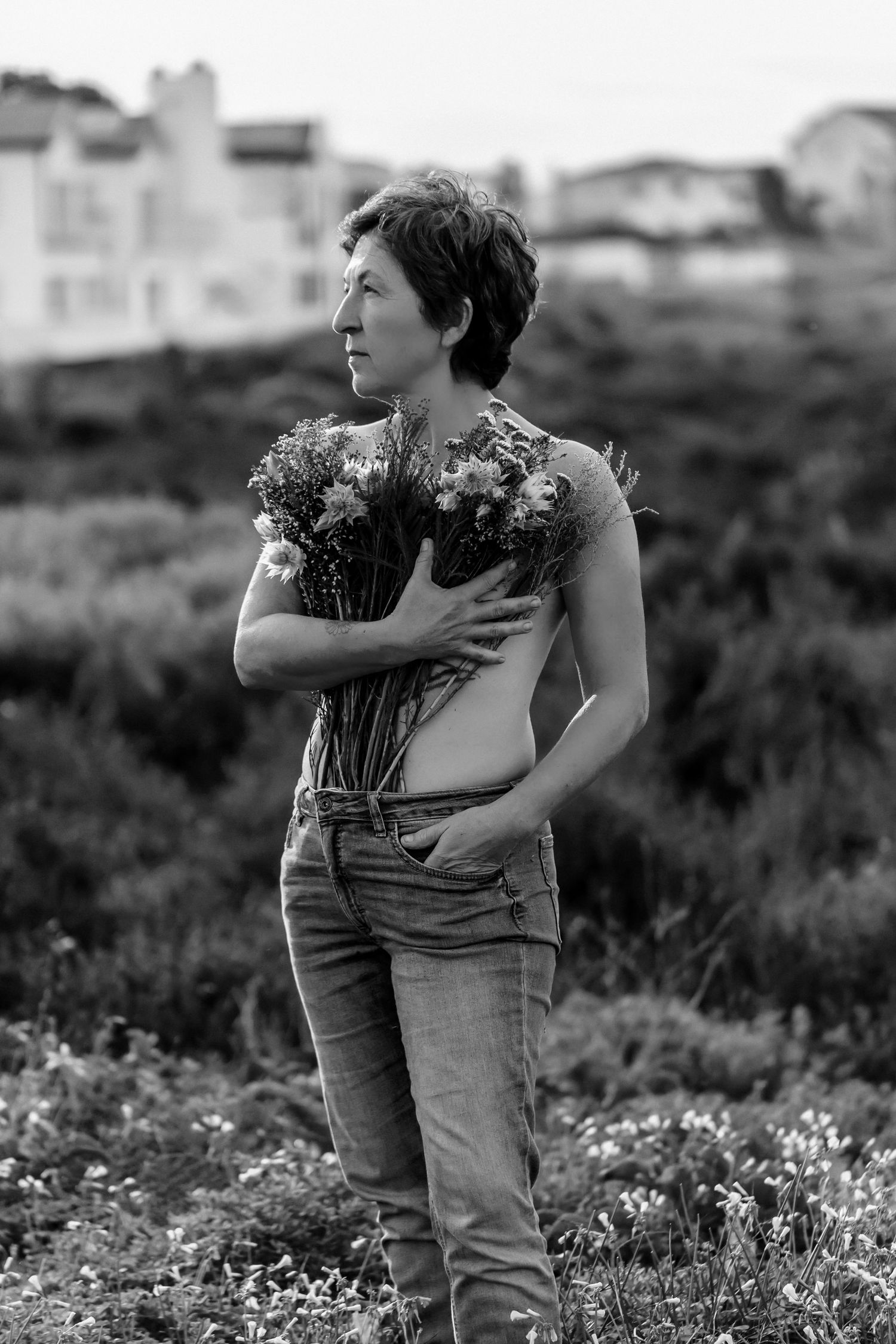 Two people embrace in a field of wildflowers against a blurred landscape in black and white.