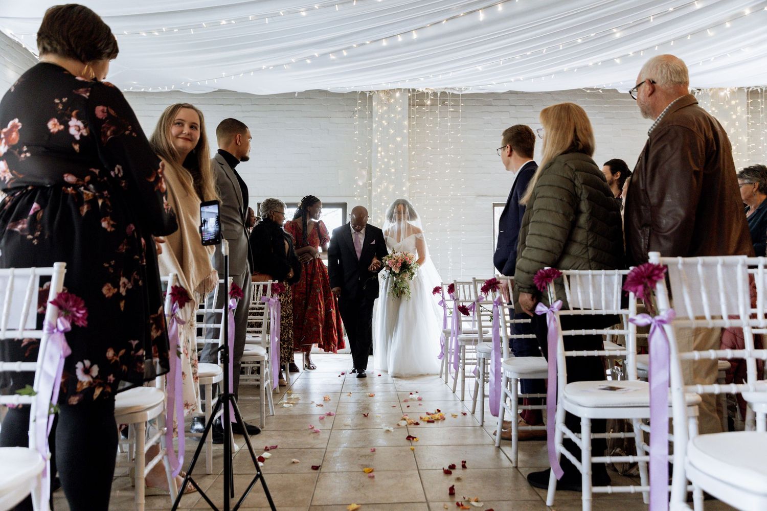 Indoor wedding ceremony in a white venue with elegantly decorated white chairs and pink accents along the aisle.