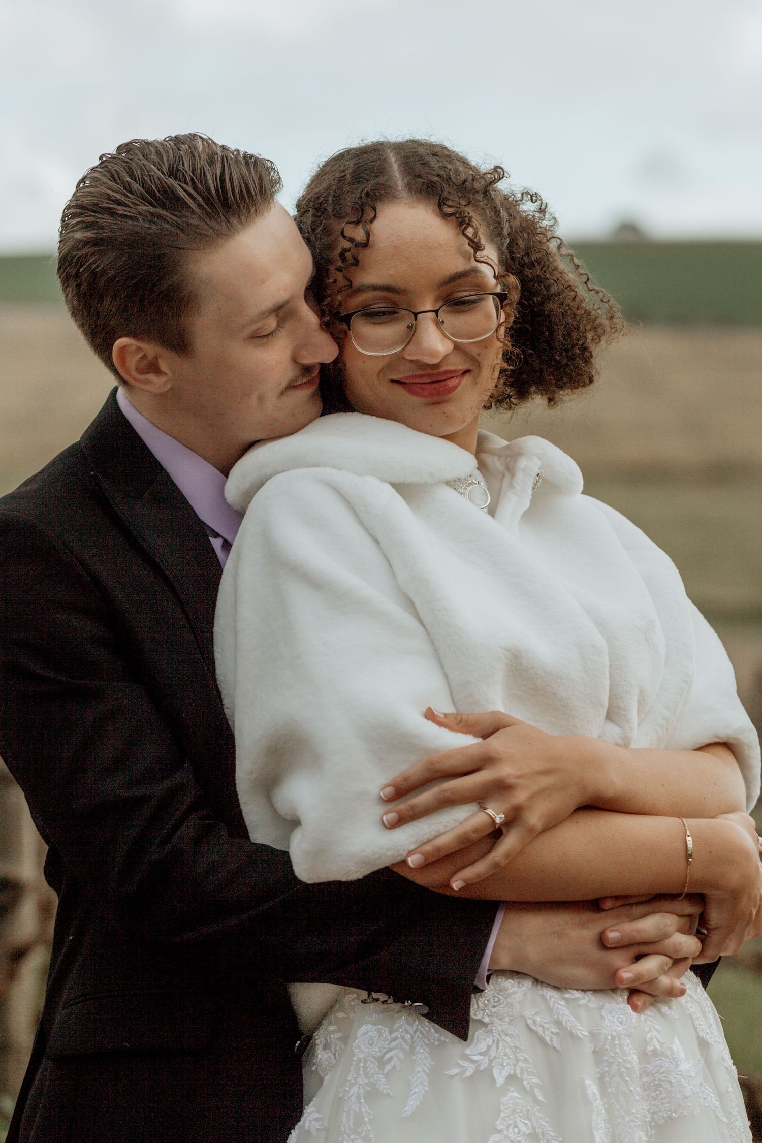 Romantic wedding couple embraces outdoors as bride wears elegant white dress and cozy wrap shawl.