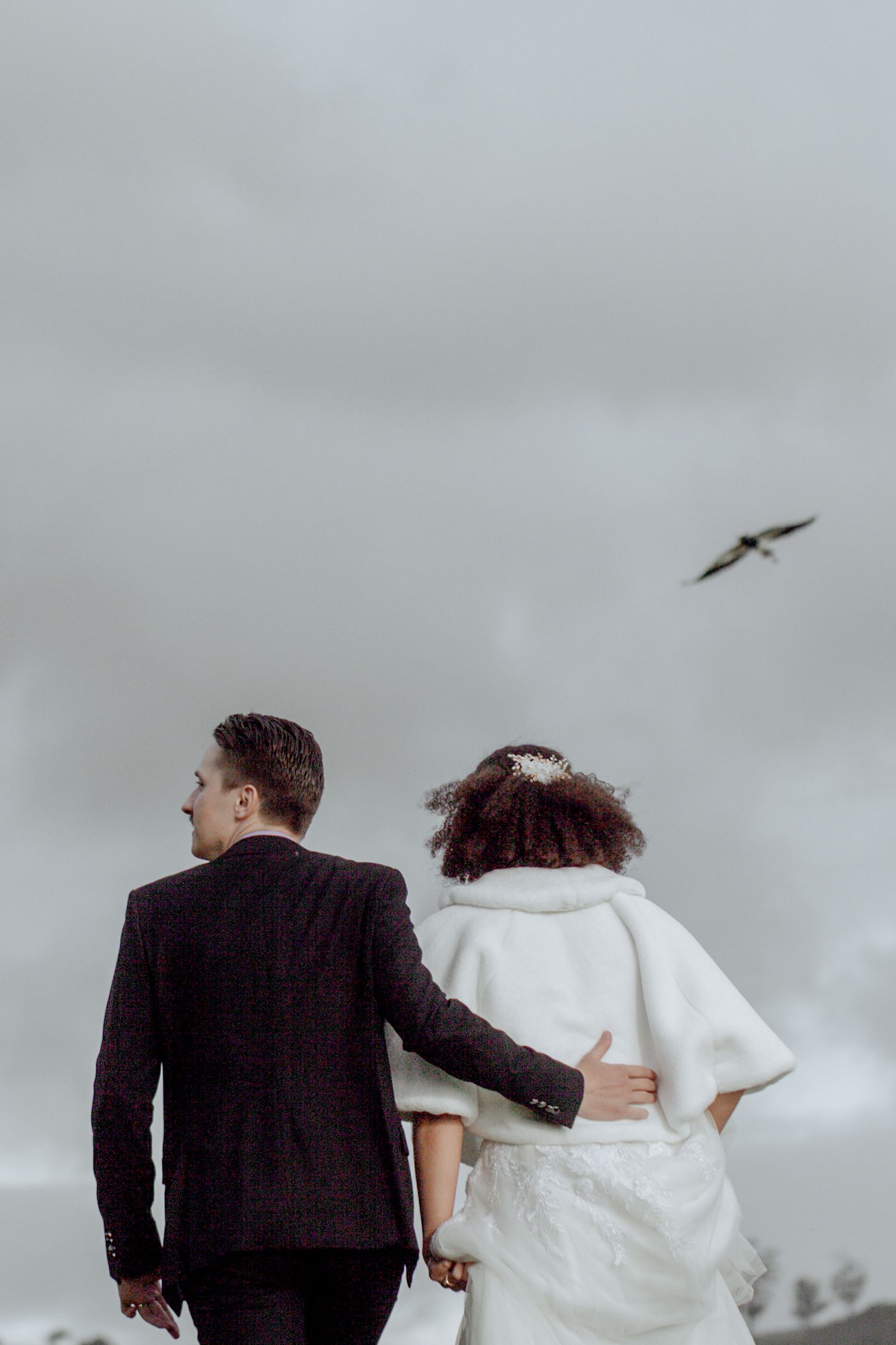 A couple in wedding attire walks together along a path as a bird flies overhead in the cloudy sky.