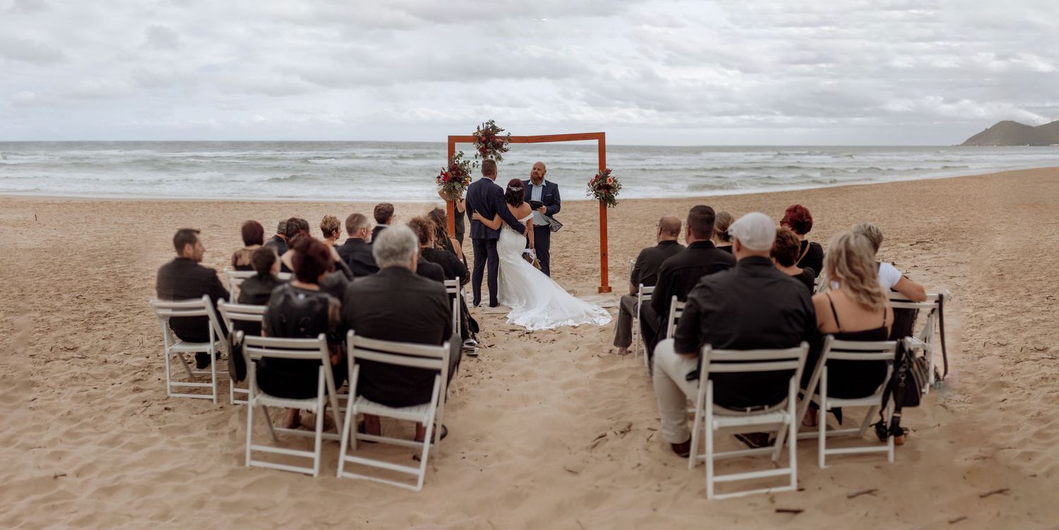 A small intimate beach wedding ceremony with wooden arch and chairs set up on the sand during an overcast day.