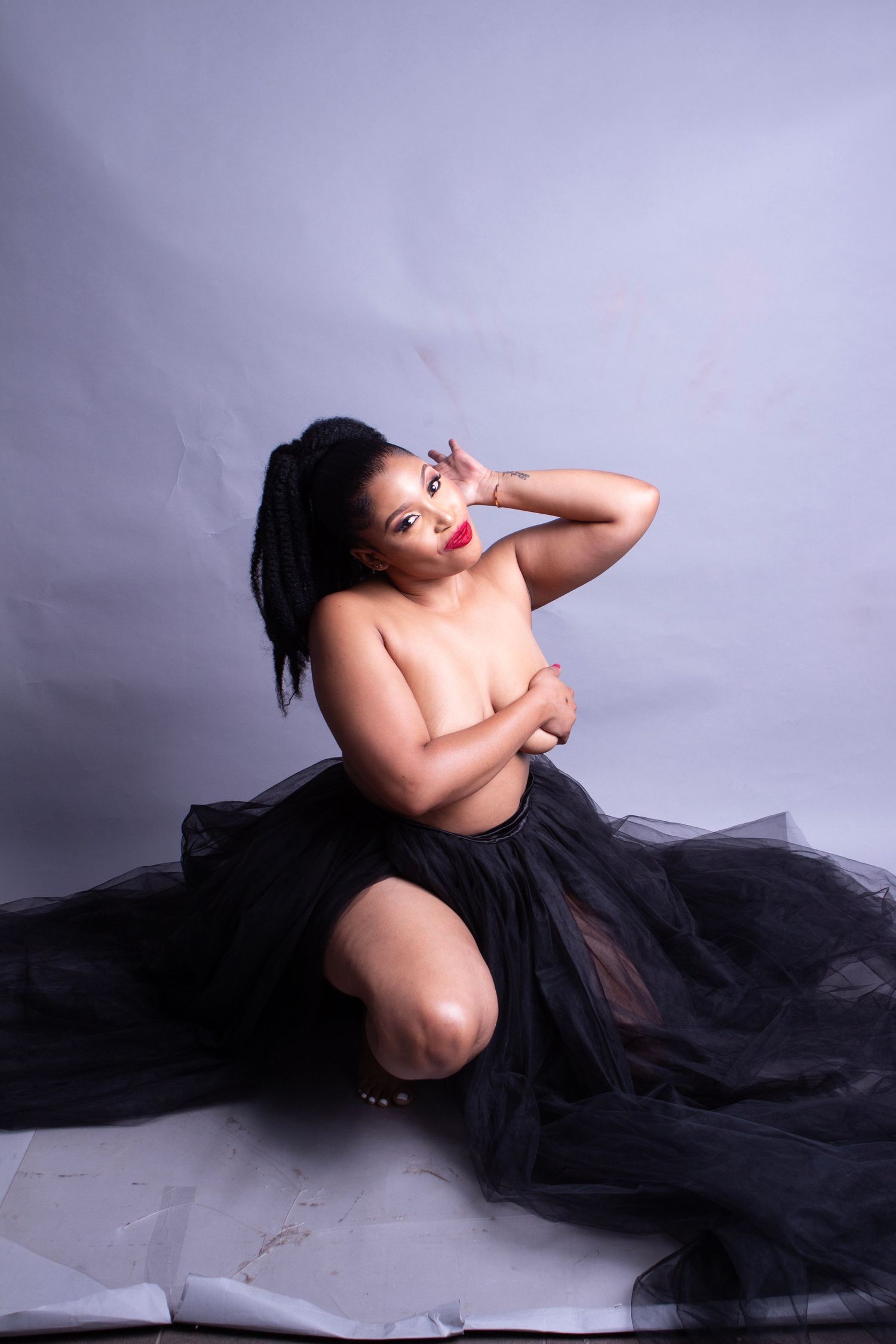 Artistic boudoir portrait featuring a figure in black tulle skirt posed against light backdrop in studio setting.