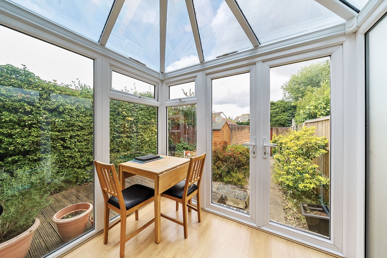 A bright sunroom or conservatory with wooden furniture and glass ceiling panels offering views of surrounding greenery.