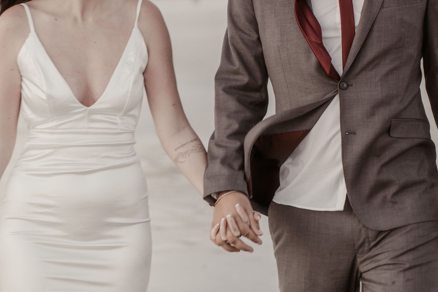 Couple holding hands in wedding attire, bride in white dress and groom in gray suit with red tie.