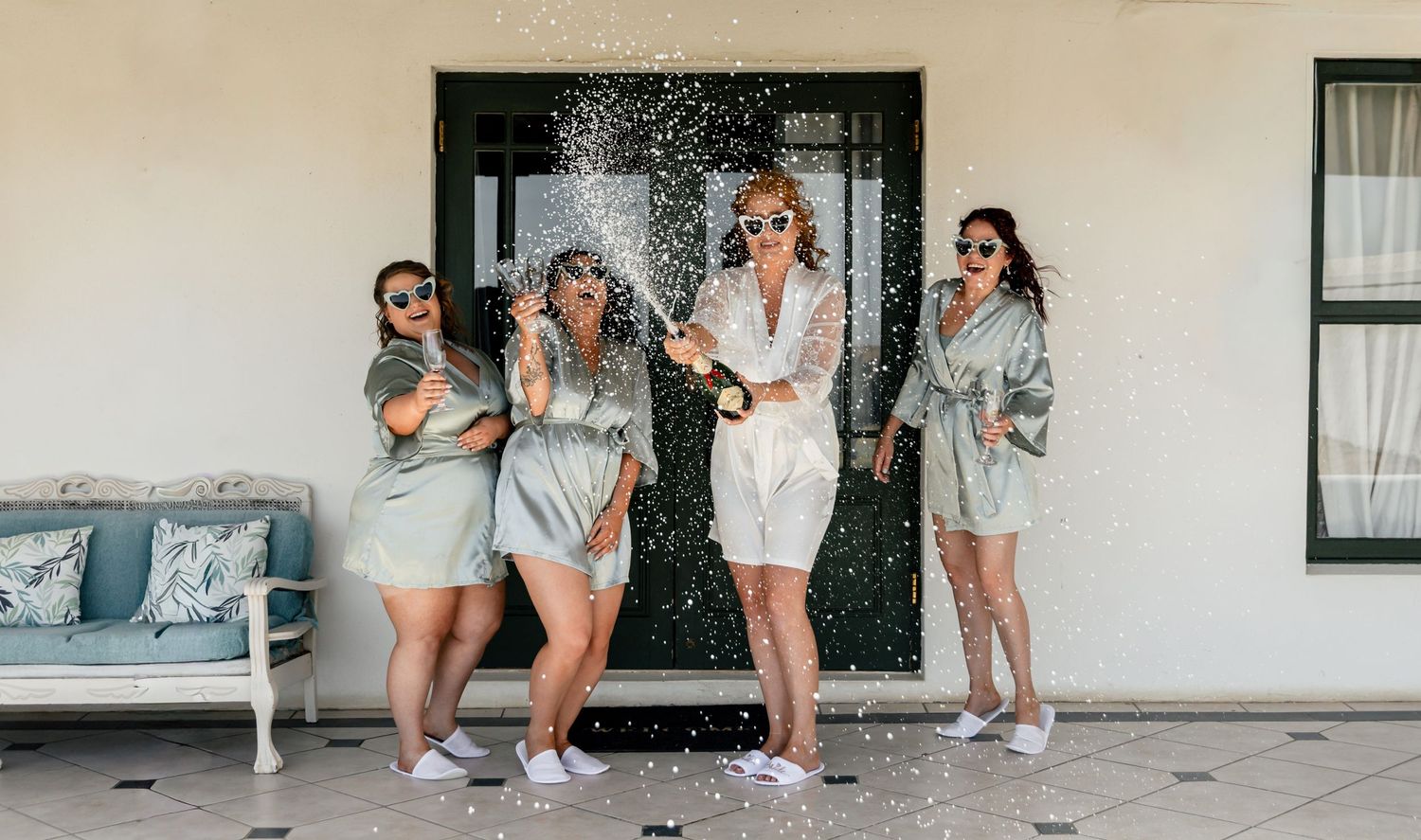Four women in matching robes celebrate with champagne spray on a sunny patio during a bridal party getting ready.