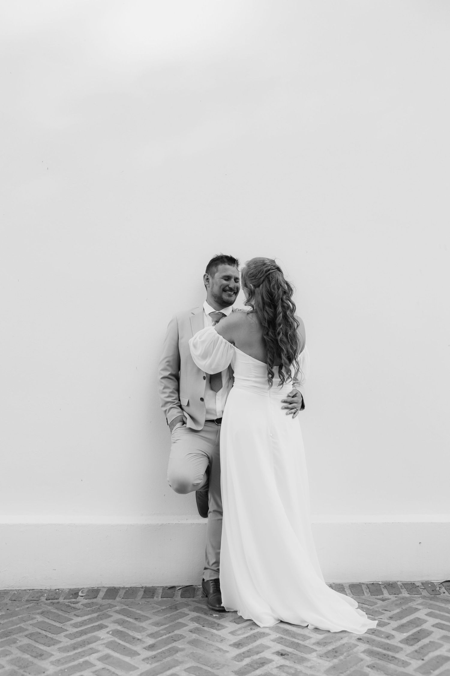 Black and white photo of a bride and groom embracing against a white wall on their wedding day.