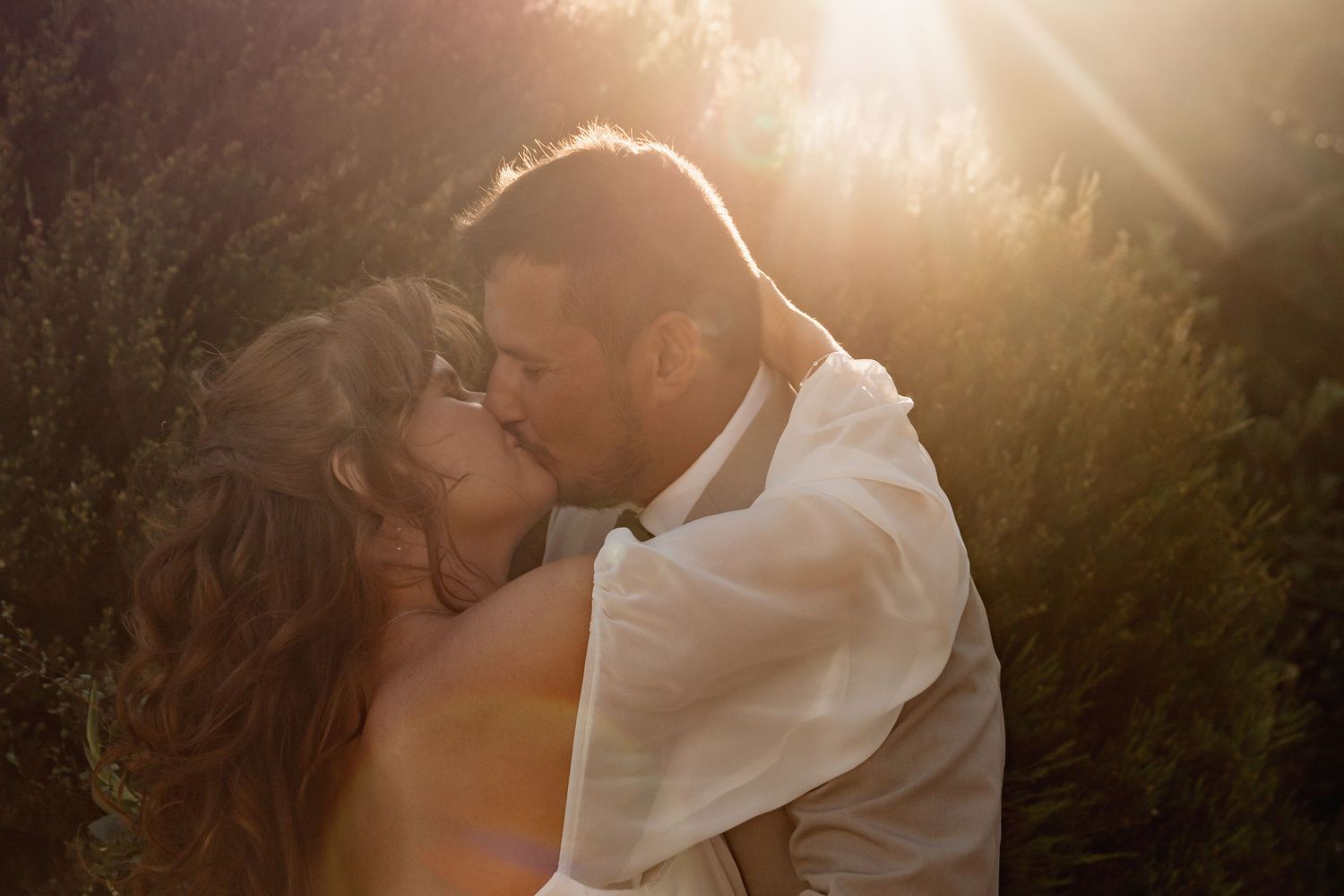 Couple sharing a romantic kiss bathed in golden sunlight with lush greenery in the background at their wedding.