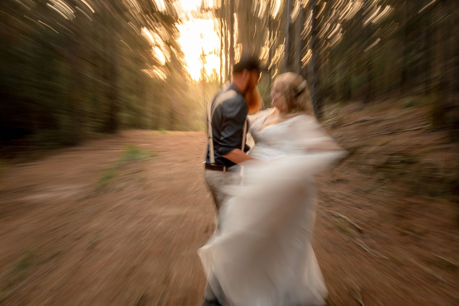 A couple in wedding attire walks through a tree-lined path during golden hour creating a romantic motion blur effect.