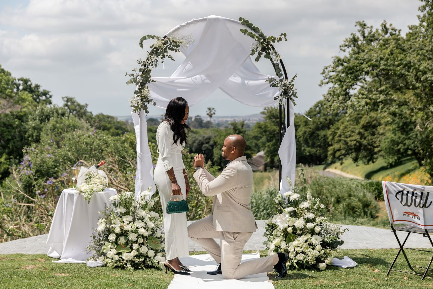 A romantic proposal takes place under a floral arch decorated with white flowers and fabric in an outdoor garden setting.