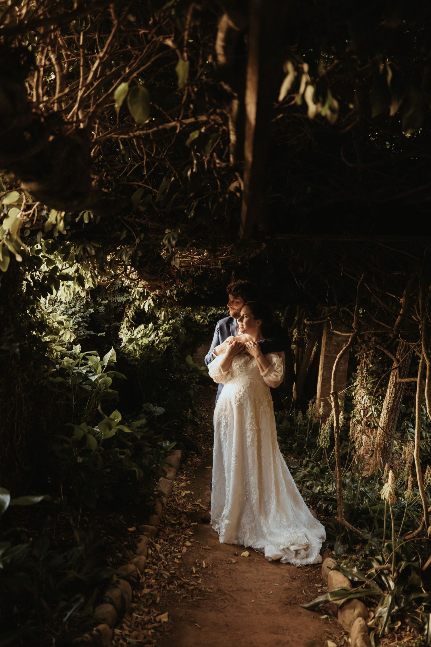 A person in a flowing white lace dress stands on a woodland path illuminated by dramatic natural lighting.