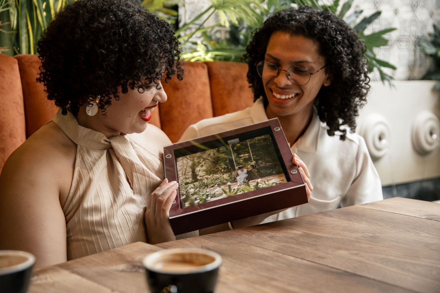 Two women smiling and sharing photos on a tablet at a cafe table with coffee cups nearby.
