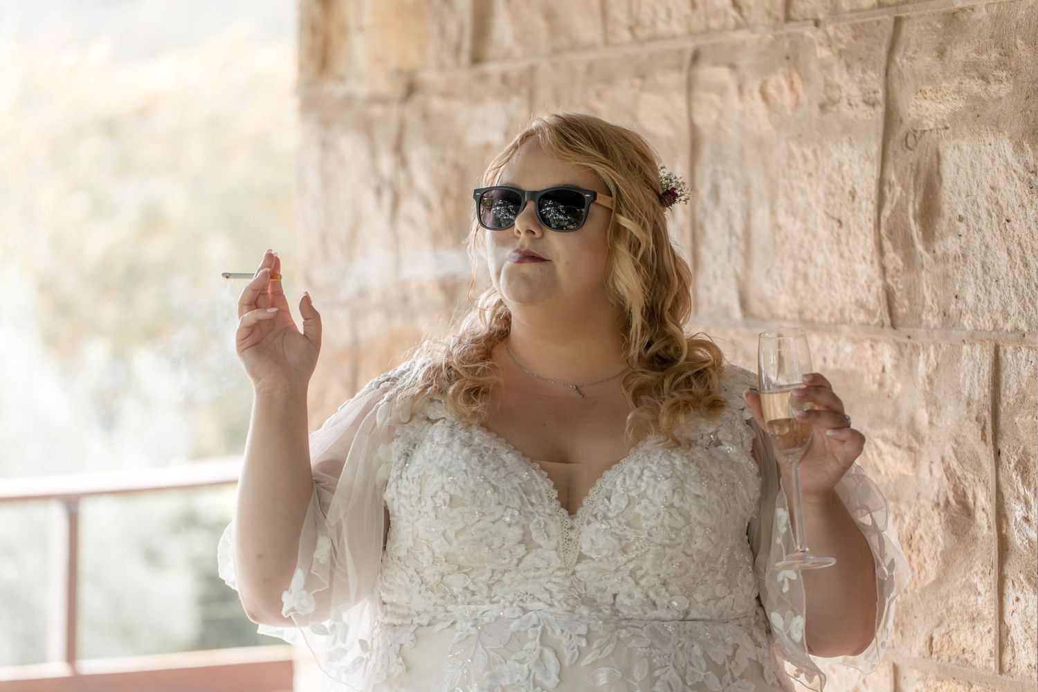 A plus-size bride in a white lace gown and sunglasses smoking a cigarette outdoors near a stone wall.