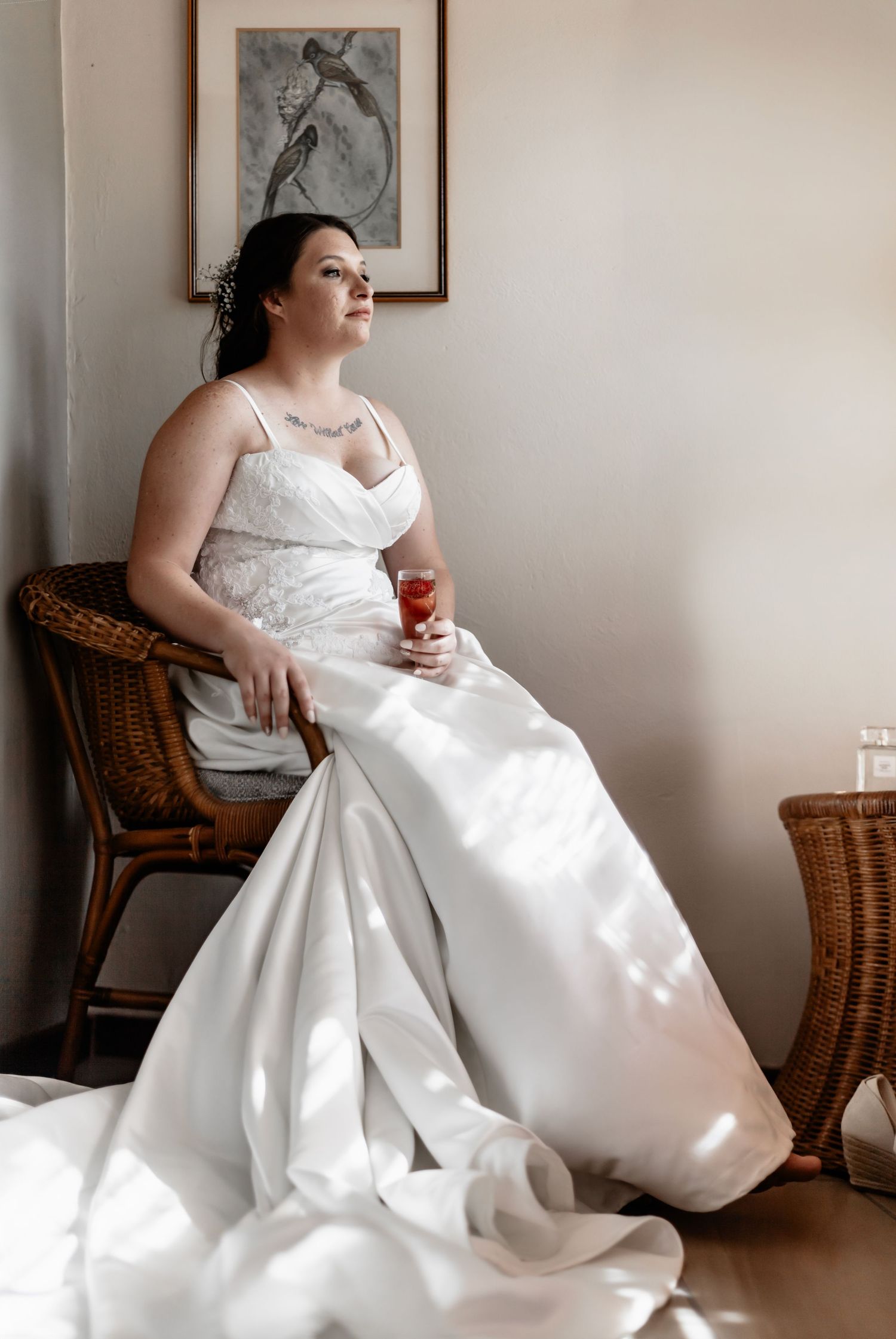 A figure in a flowing white wedding dress relaxes gracefully in a wicker chair next to a sunlit window.