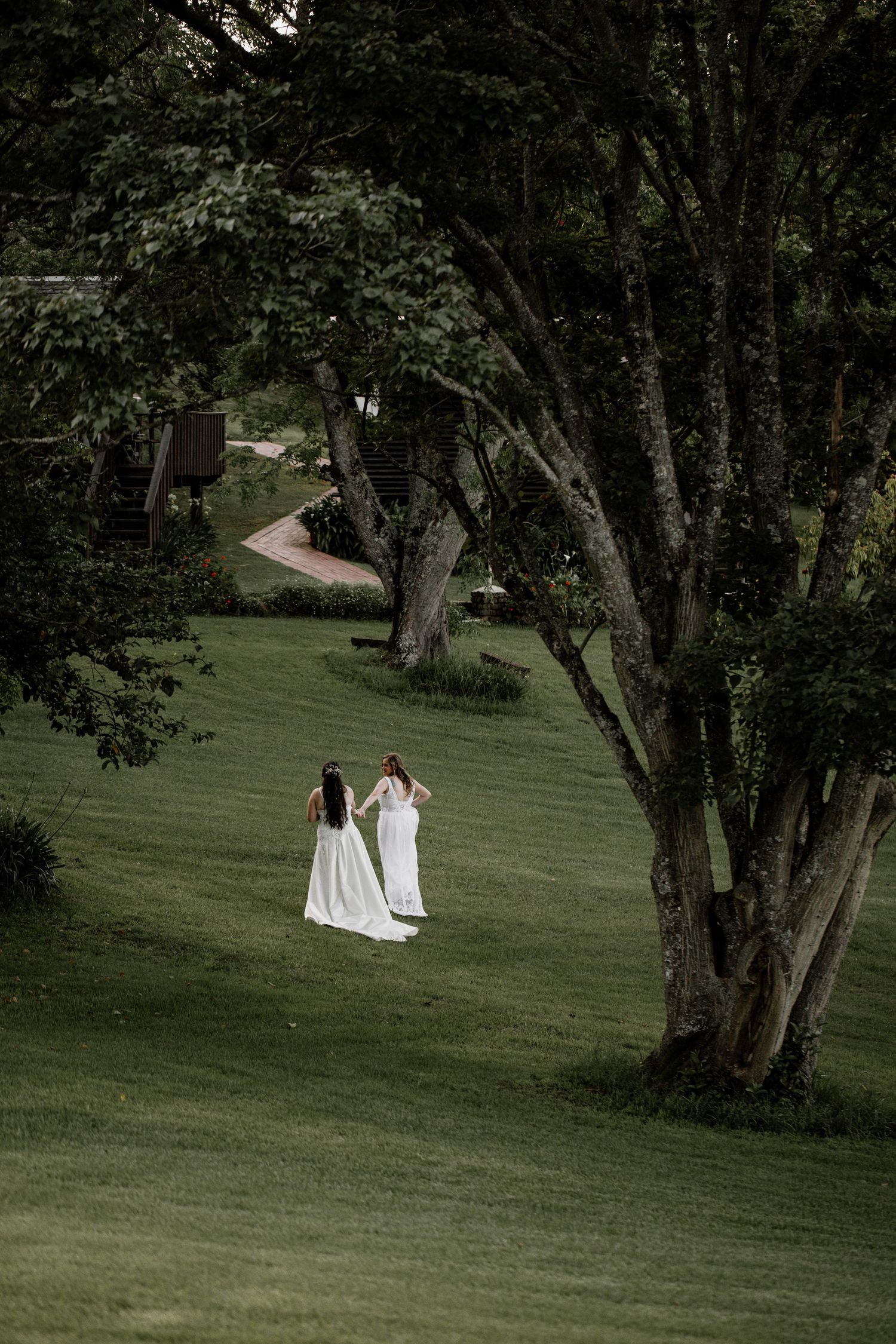 Two figures in flowing white dresses walk together on a lush green lawn surrounded by tall trees in a moody garden setting.