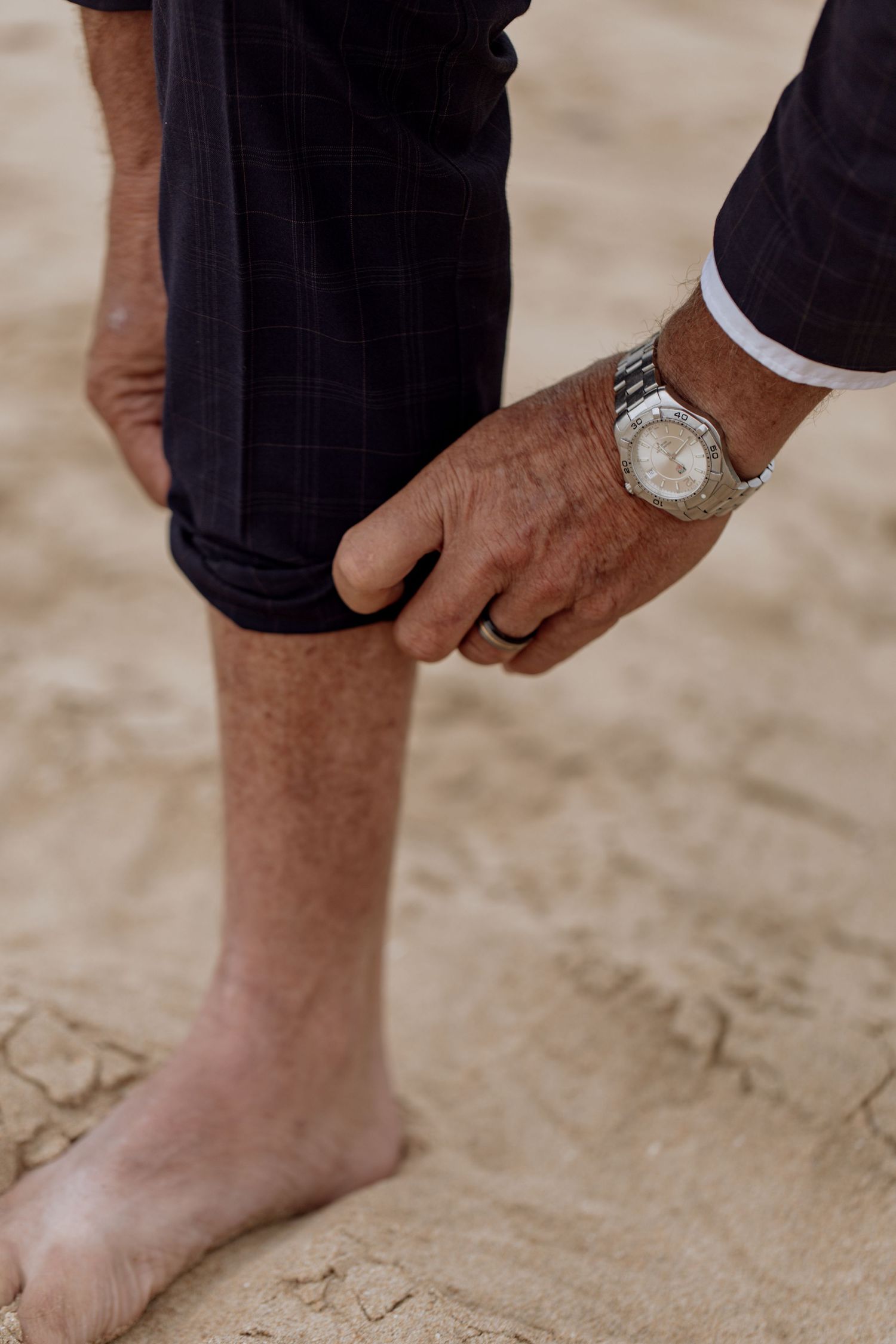 Close-up of a hand wearing a silver watch and ring adjusting dark pants leg while standing barefoot on sandy ground.