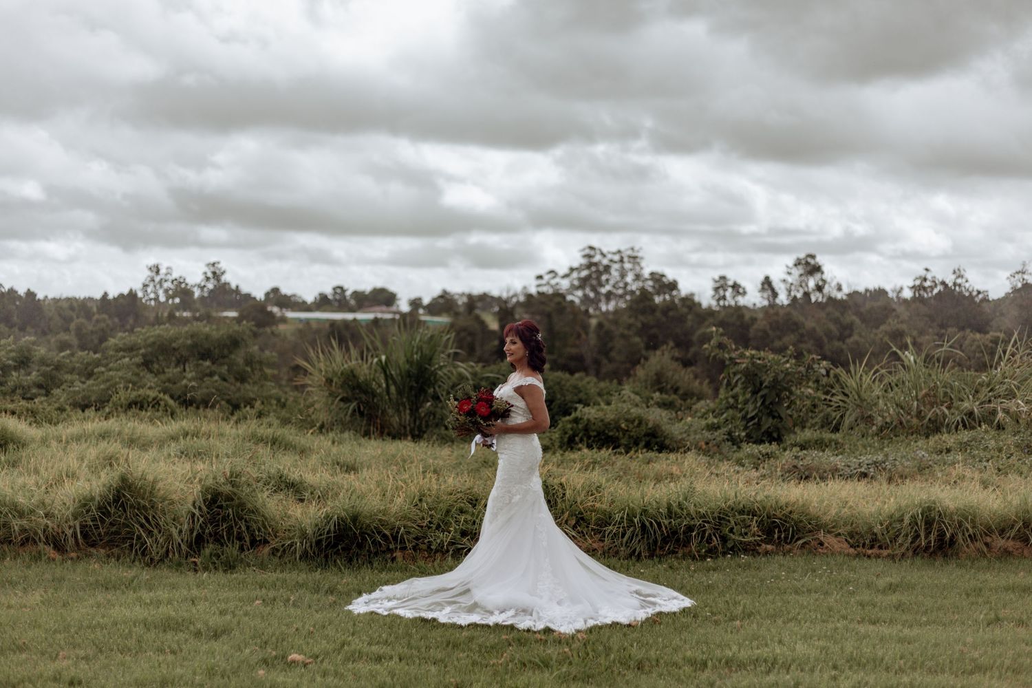 A white lace wedding dress with a long train flows across green grass under dramatic cloudy skies.