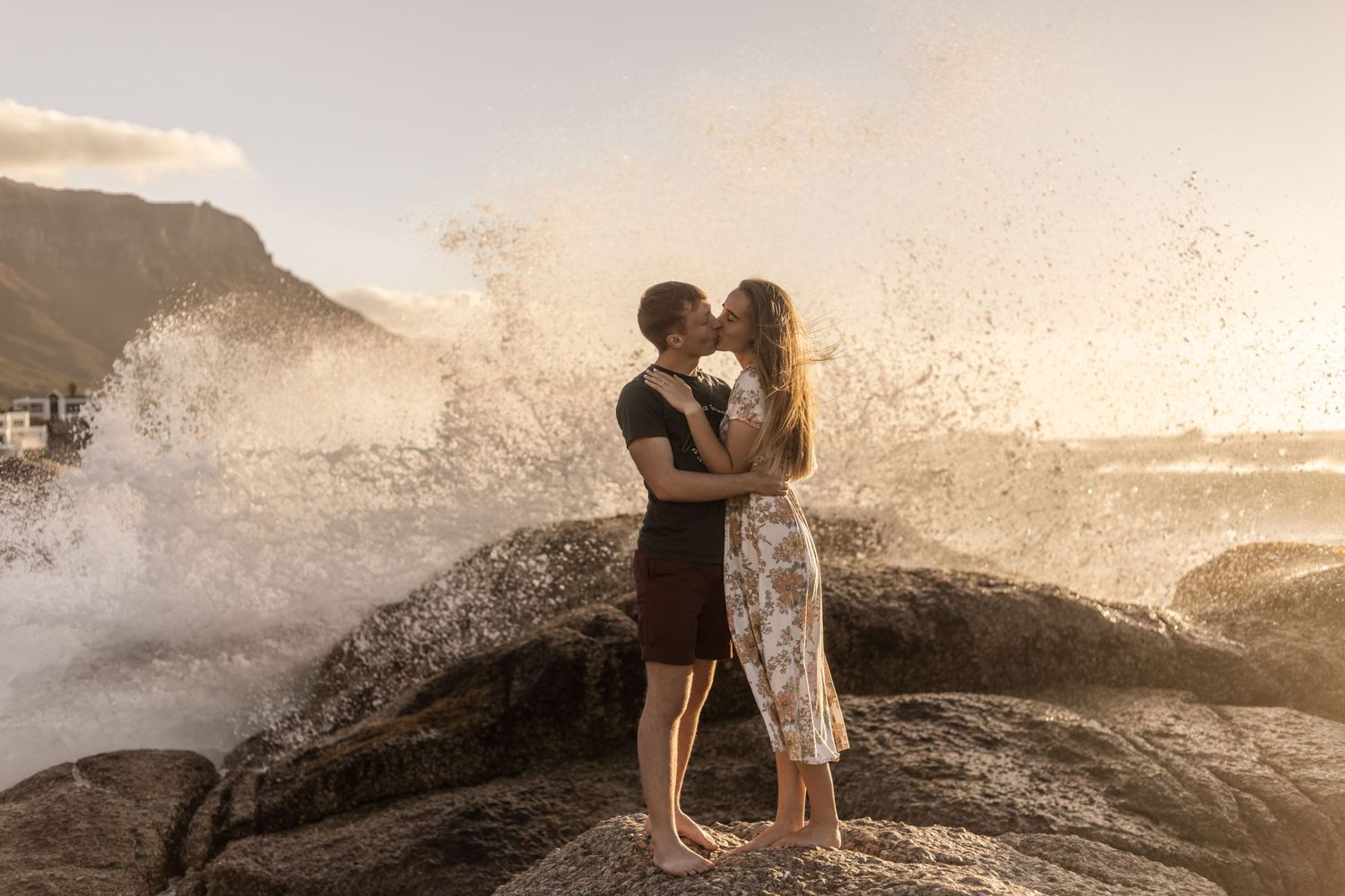 Two silhouetted figures embrace on rocky shore as ocean waves crash dramatically at sunset creating a romantic scene.