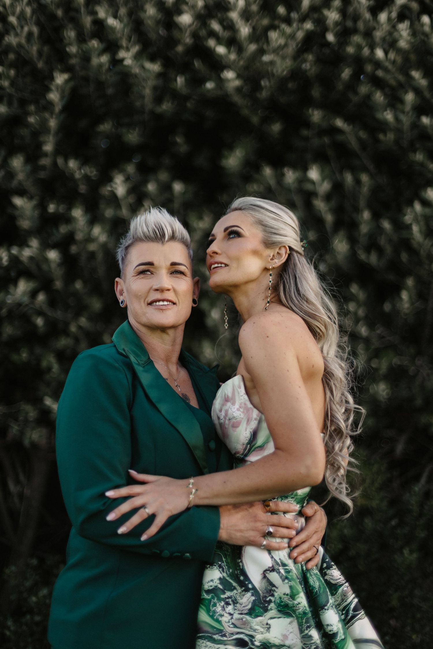 Two people share joyful laughter and embrace while wearing green and floral outfits against a natural backdrop.
