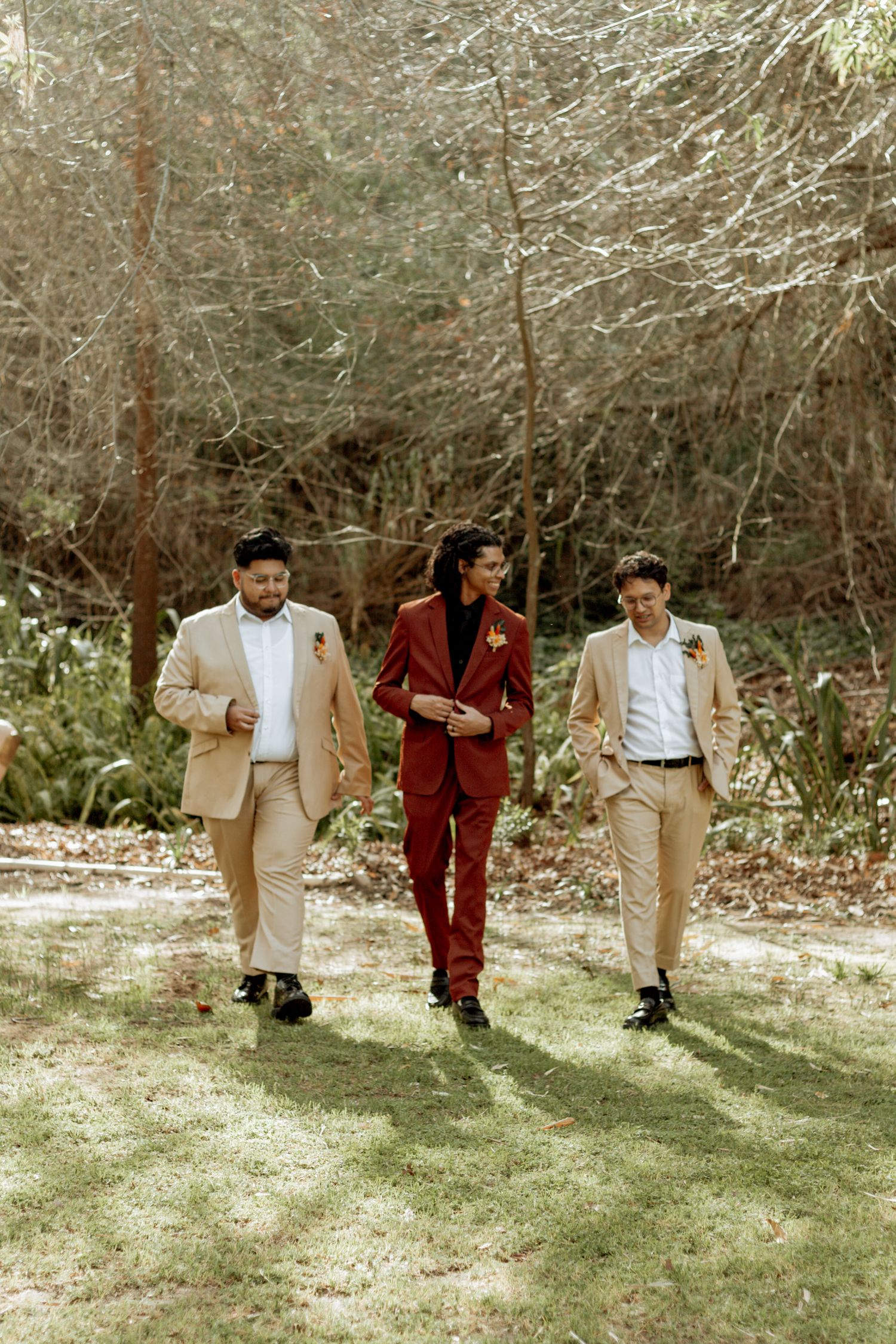 Three people in coordinating tan and burgundy formal attire walk together in a wooded outdoor setting.