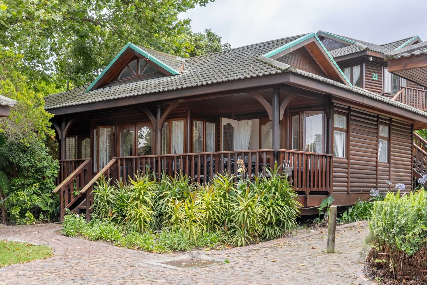 Rustic brown wooden cottage with wraparound porch and lush garden landscaping features tropical plants and stone path.