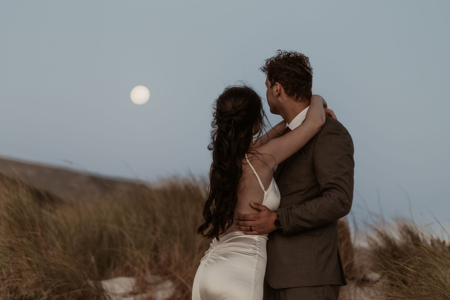 Couple embracing on sandy dunes under a full moon at dusk, bride in white gown and groom in brown suit.