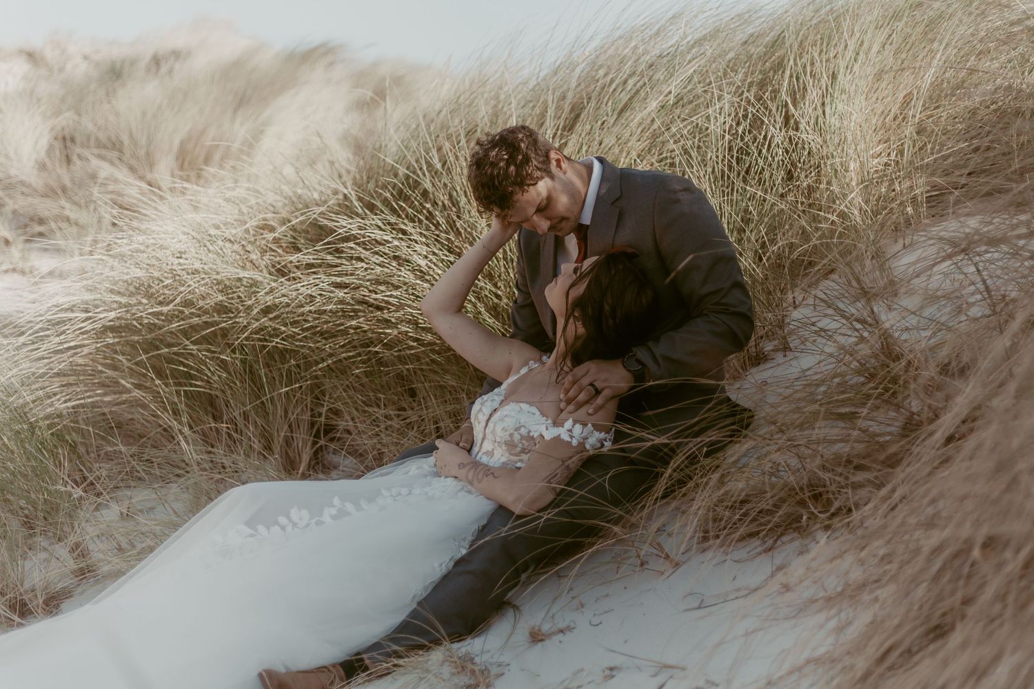 Couple in wedding attire sitting in sand dunes surrounded by tall grass during romantic outdoor elopement photoshoot.