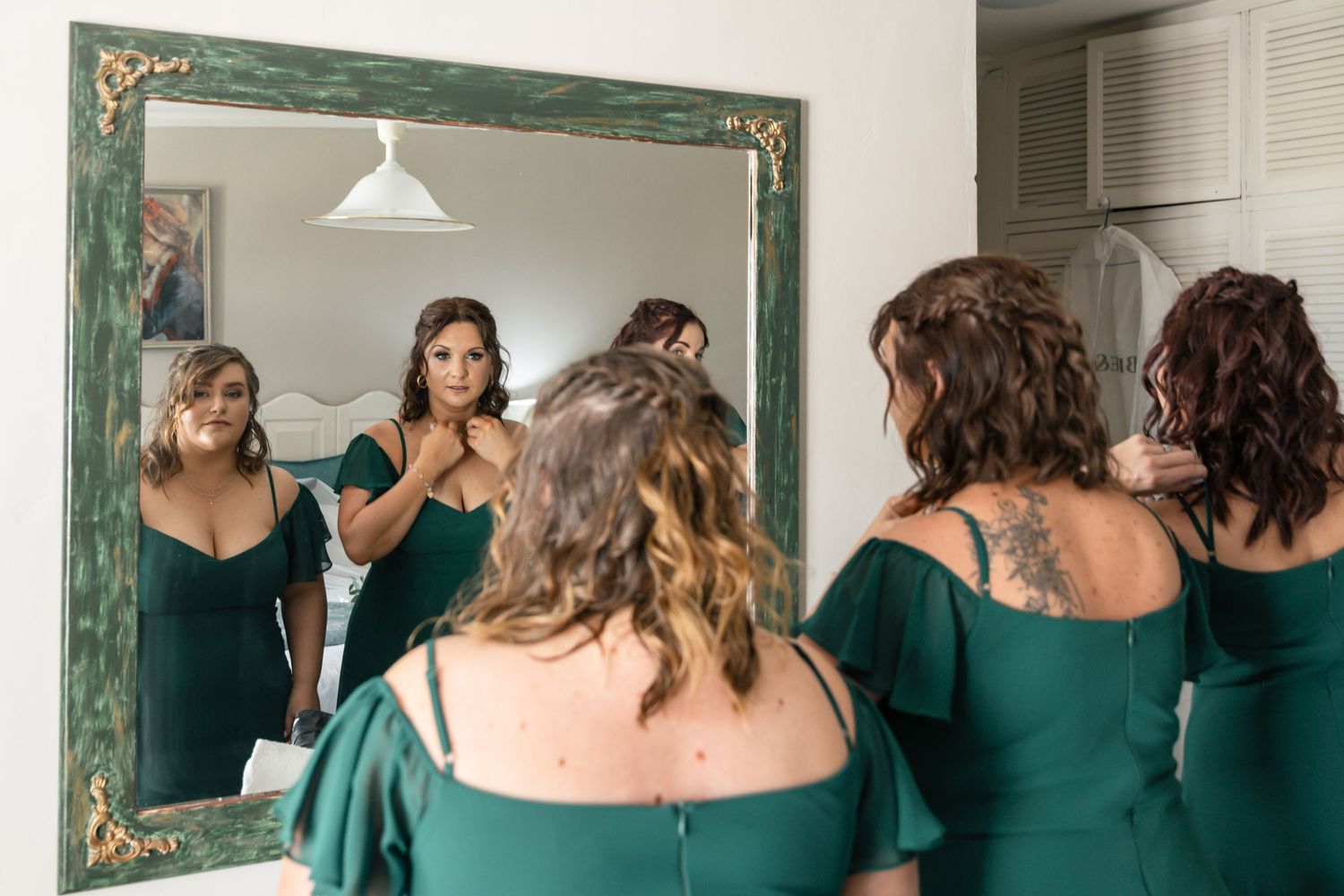 Bridesmaids in green dresses help each other get ready, reflected in an ornate green-framed mirror.
