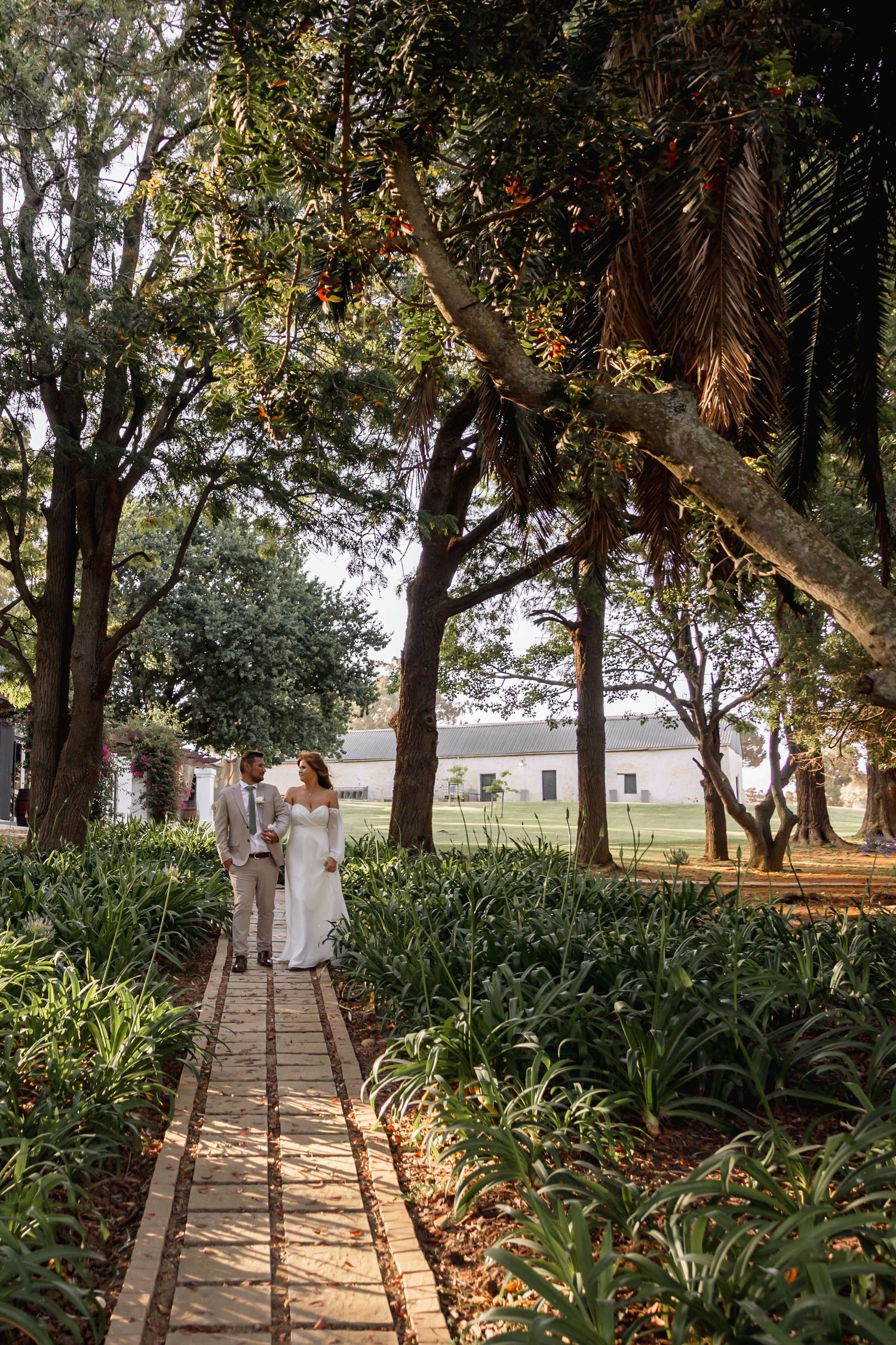A bride and groom walk hand in hand along a brick garden path surrounded by lush greenery and tall trees.