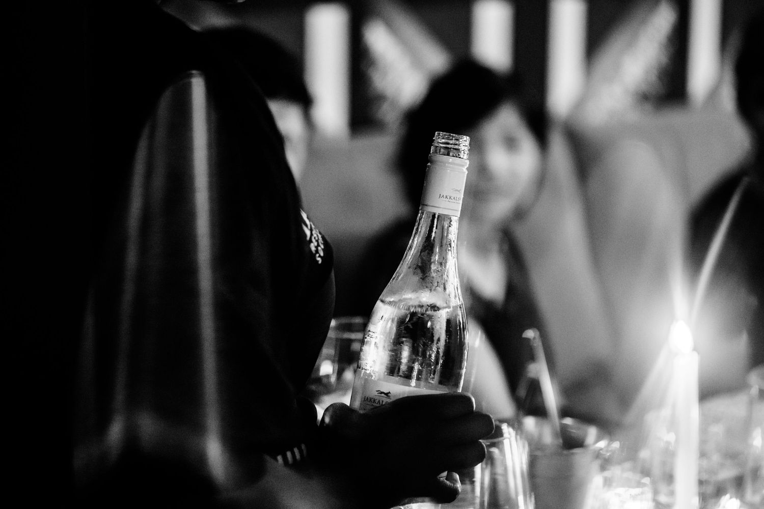 A black and white image of a vodka bottle being held up in a bar or club atmosphere against a blurred background of hands.