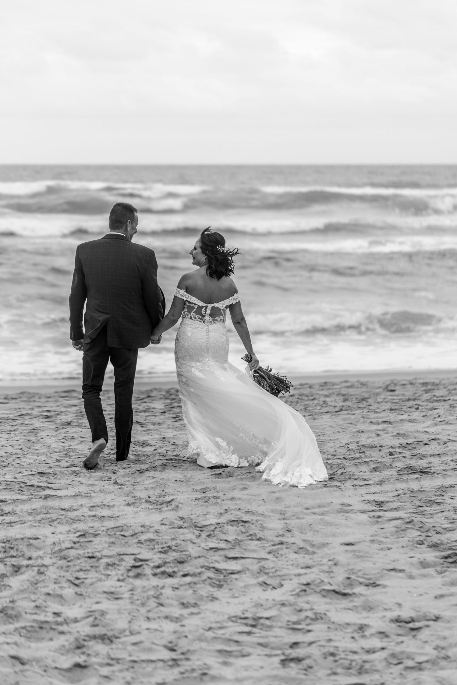 A couple walks hand in hand along a beach during their wedding photoshoot as waves crash in the background.
