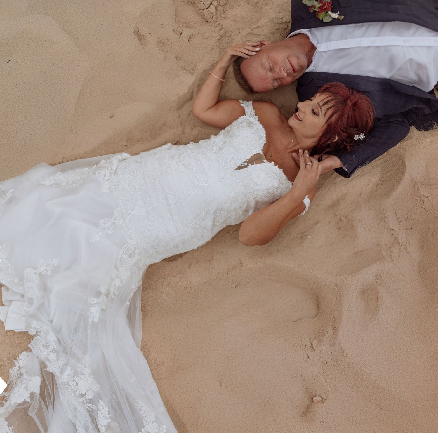A bride and groom lying on beach sand, with the bride wearing a white lace wedding dress.