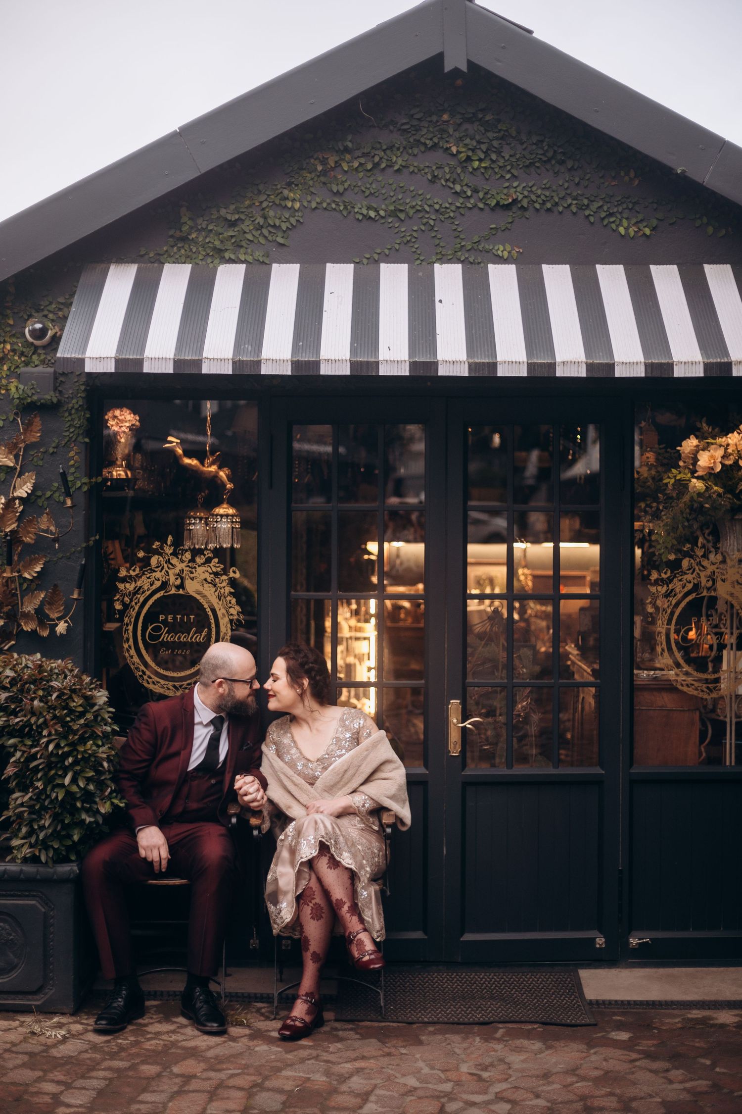 Couple sitting together outside Hotel Chocolat shop with striped awning, sharing a romantic moment.