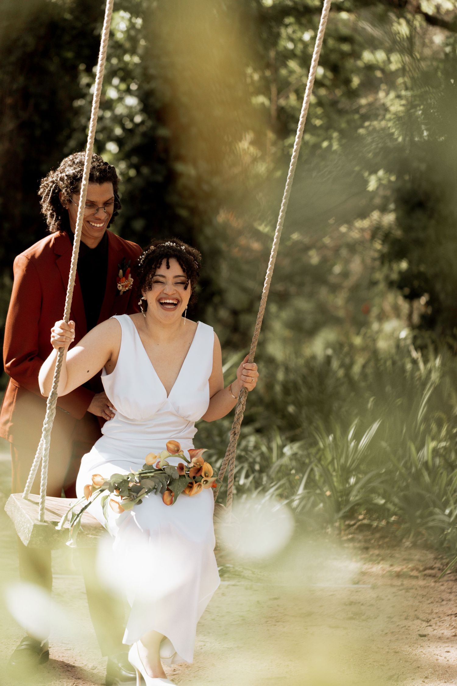 A joyful scene shows someone wearing a white dress laughing while sitting on a rustic wooden swing in a garden setting.