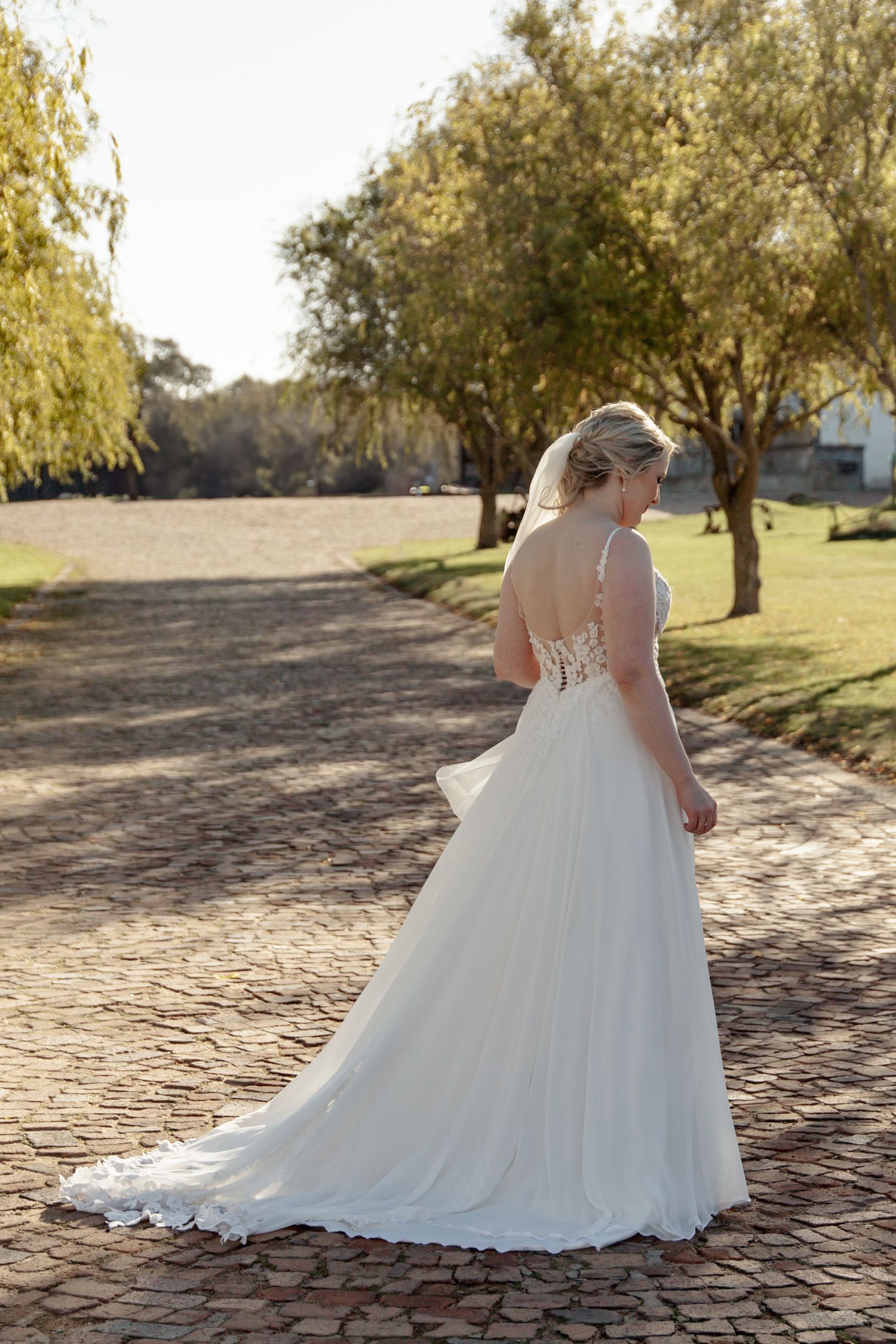 A bride in a flowing white wedding dress stands on a stone path surrounded by trees during golden hour.