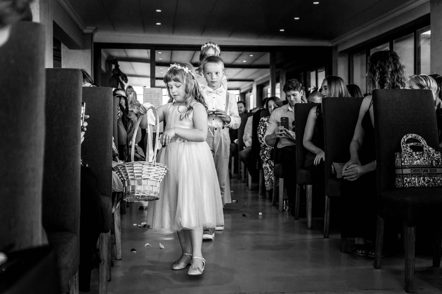 A young flower girl in a white dress walks down an aisle lined with wedding guests in black and white photography.