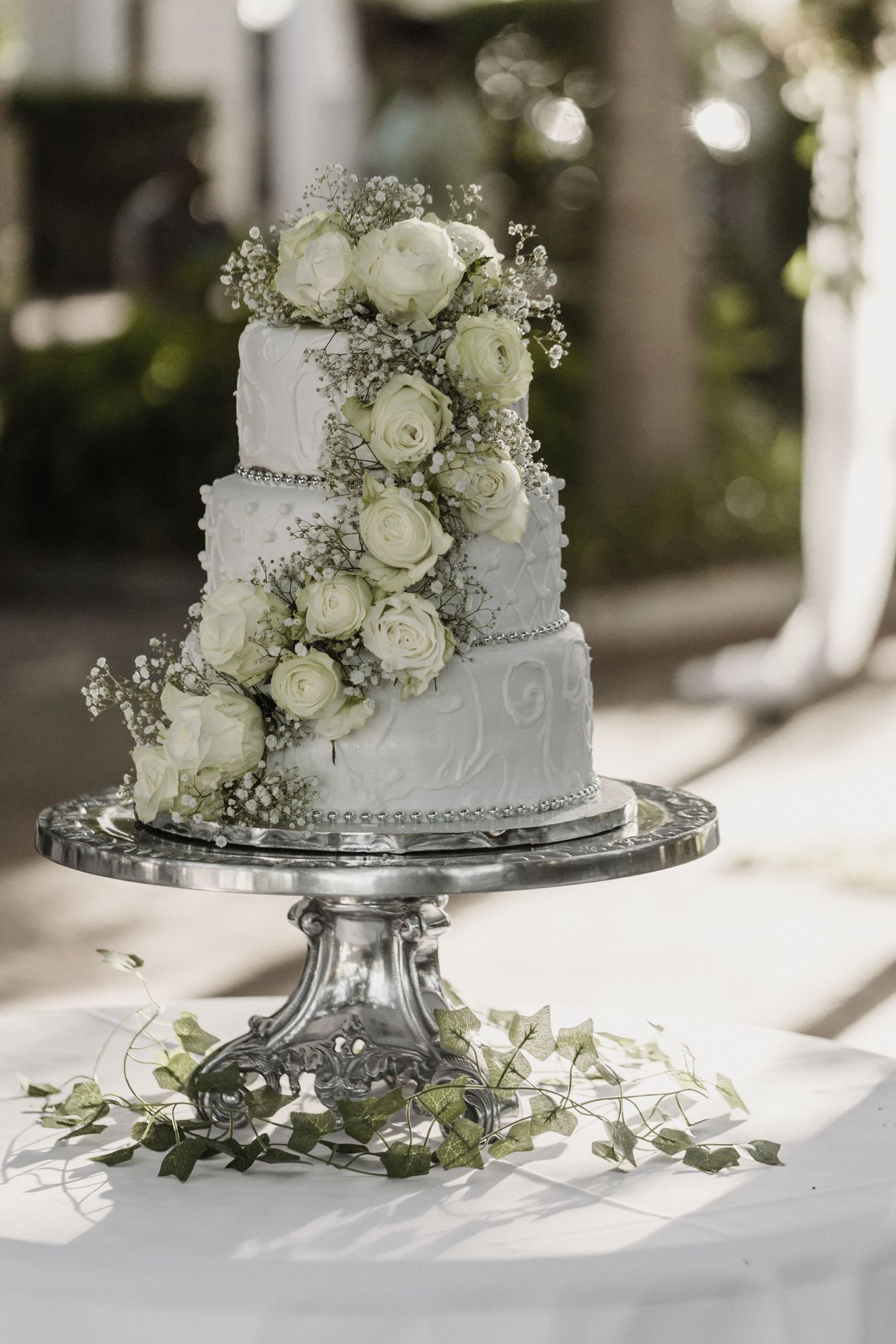 Elegant white wedding cake decorated with fresh white roses and baby's breath on silver cake stand in natural lighting.