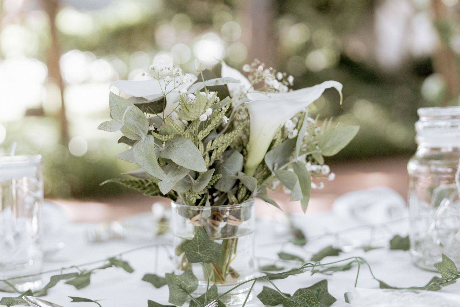 White calla lilies and greenery centerpiece arrangement on an elegant table setting with glassware.