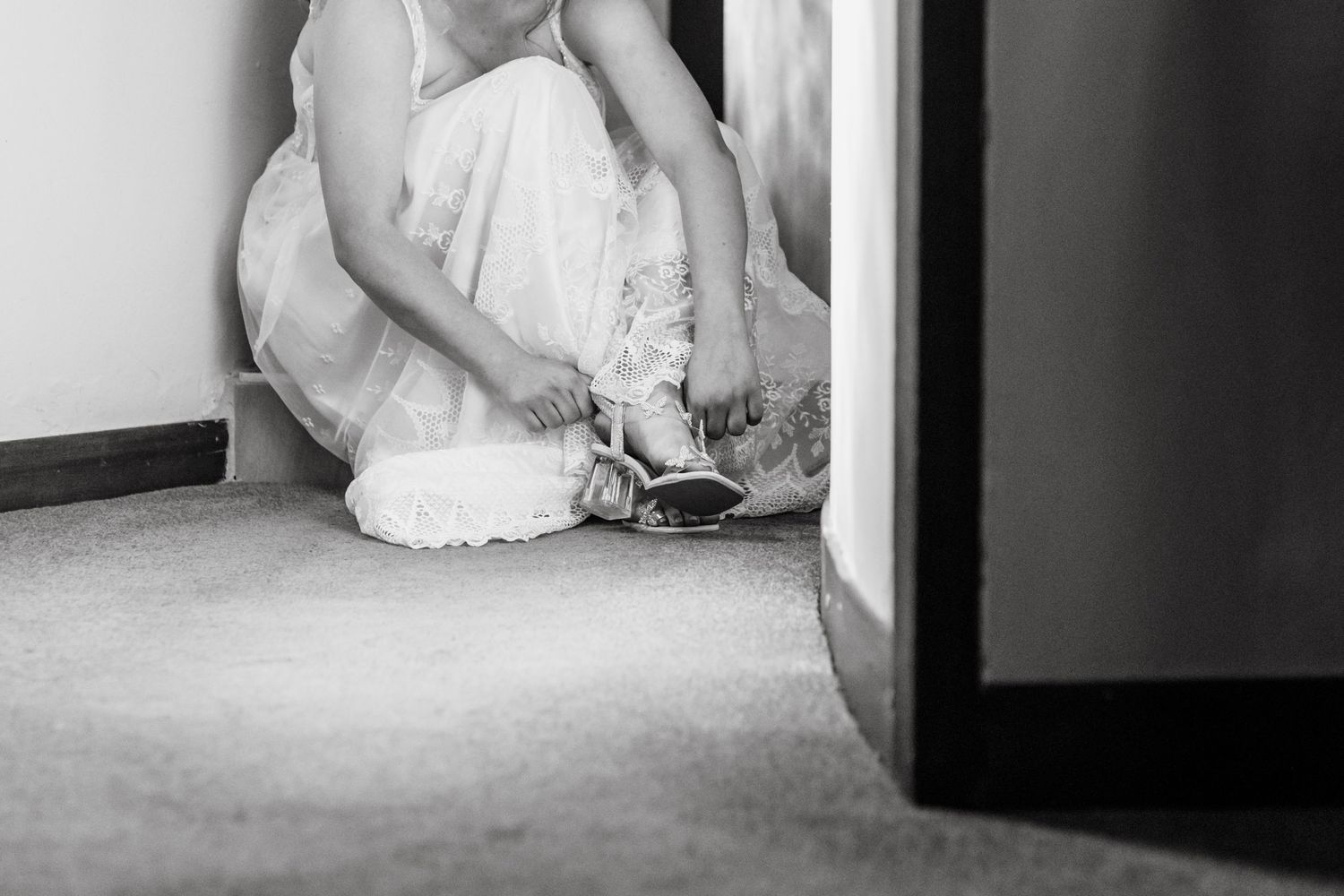 Black and white image of someone in a flowing white dress crouched down adjusting their shoe on a carpeted floor.