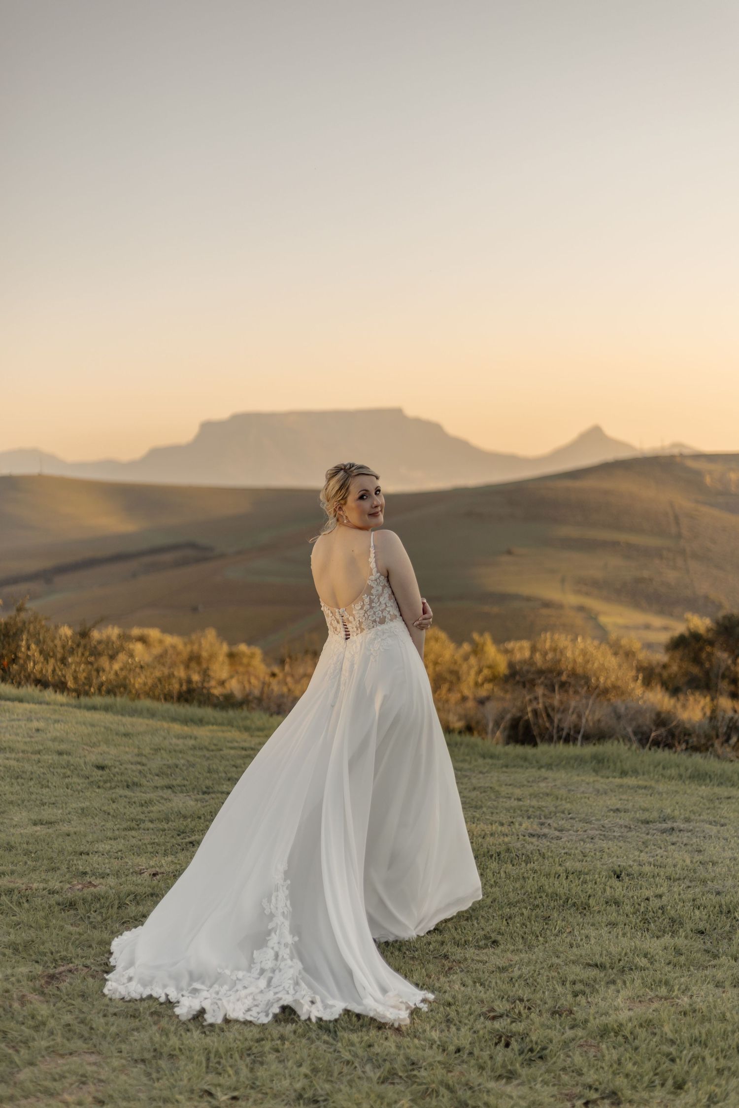 Beautiful wedding dress with flowing skirt and lace details against mountain landscape at golden hour sunset.
