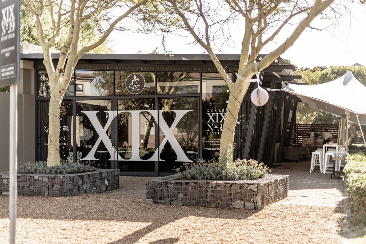 Modern industrial outdoor restaurant space with XIX signage and rustic decor under shady trees.