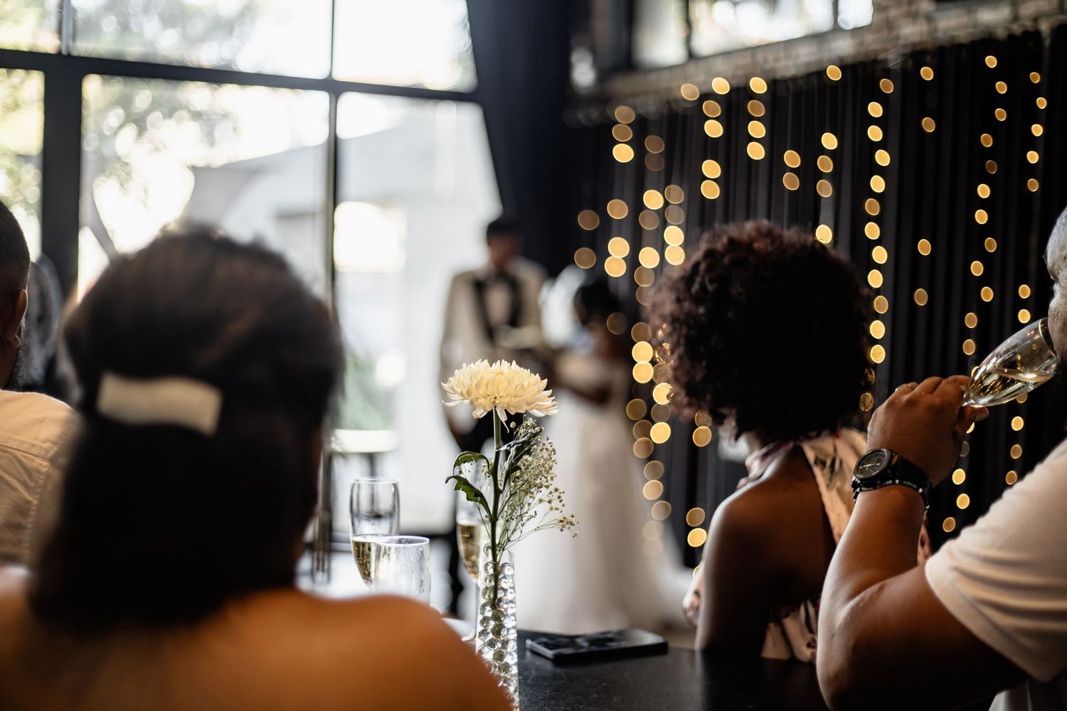 A large celebration room with hanging string lights and people seated at long tables decorated for a festive event.