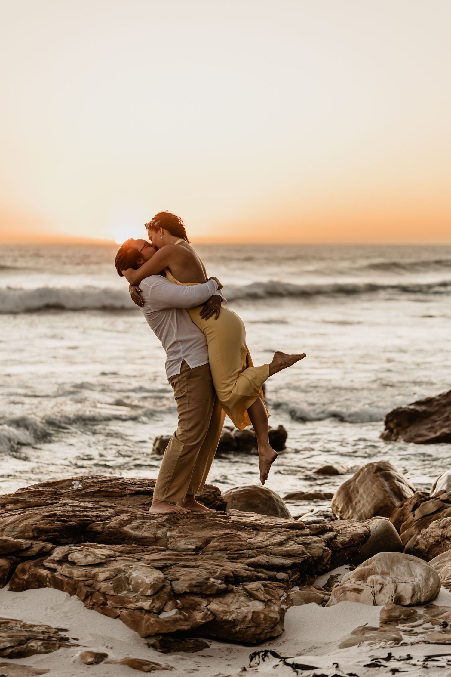 Romantic sunset beach moment with couple embracing on rocky coastal outcrop by ocean waves at golden hour.