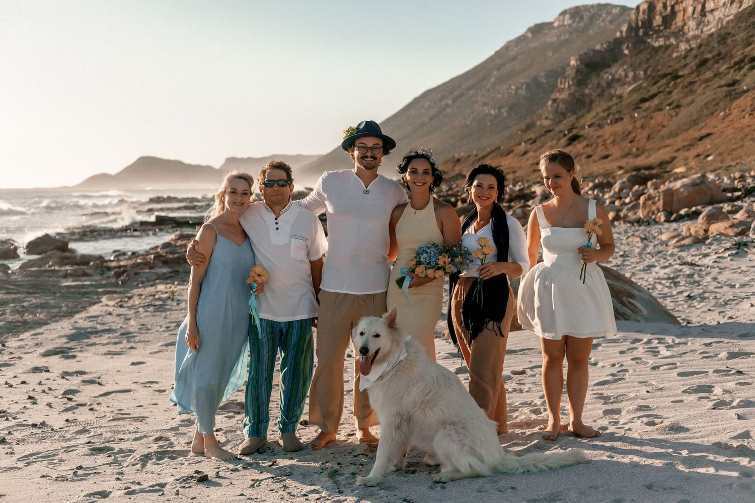 Group of people walking and posing together on a scenic rocky beach at sunset with mountains in the background.
