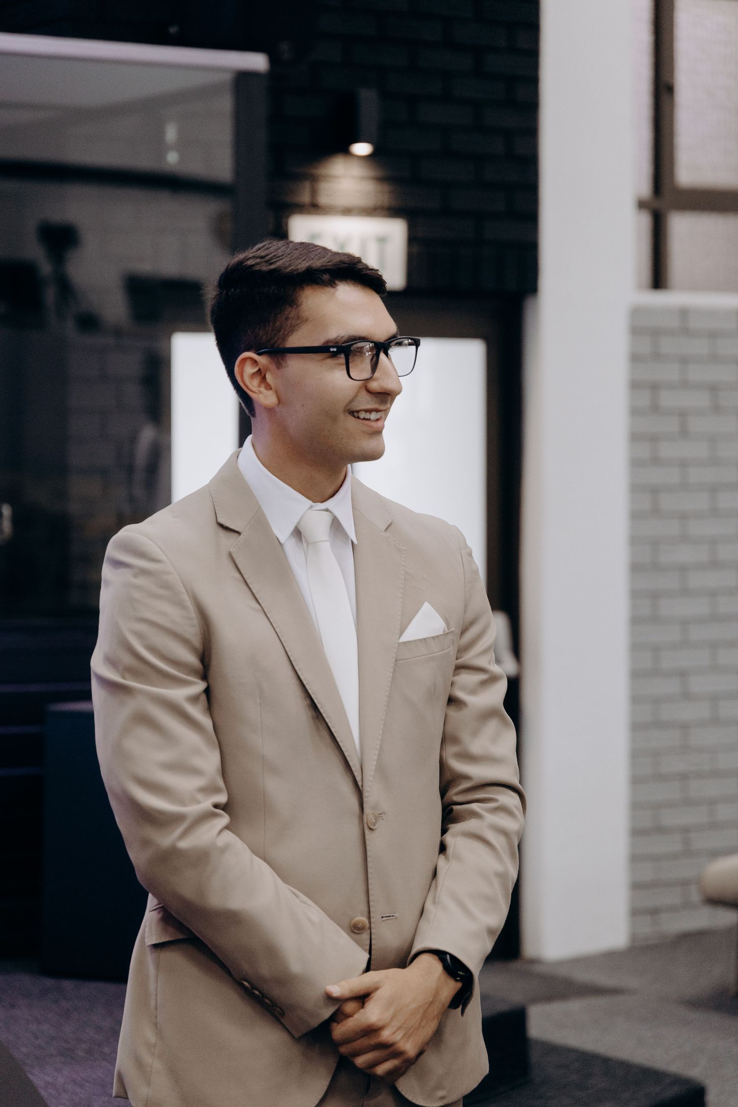 A person in a beige suit and glasses poses in front of a white brick wall in a professional setting.