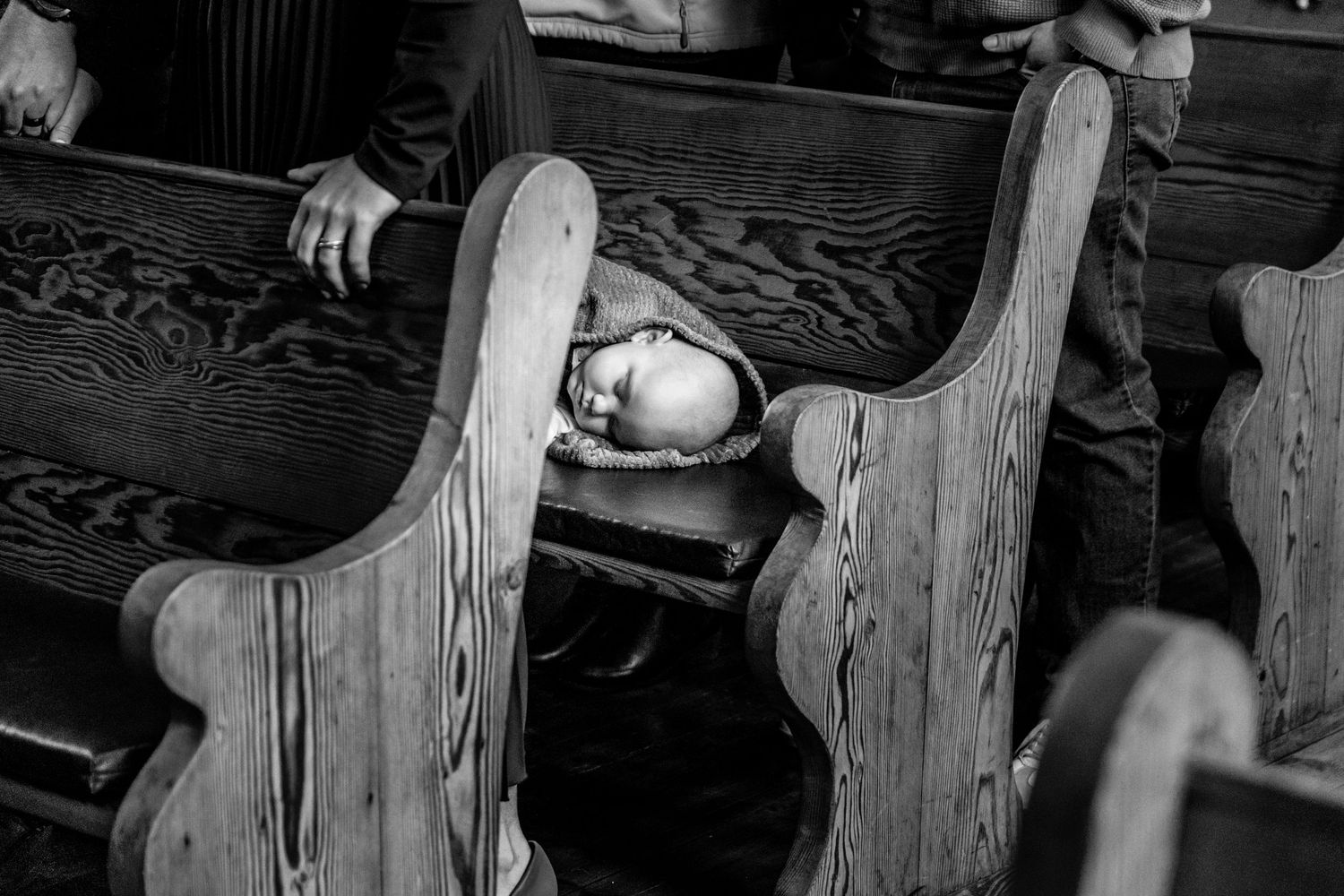Black and white photo of a sleeping baby nestled between wooden church pews during a service.