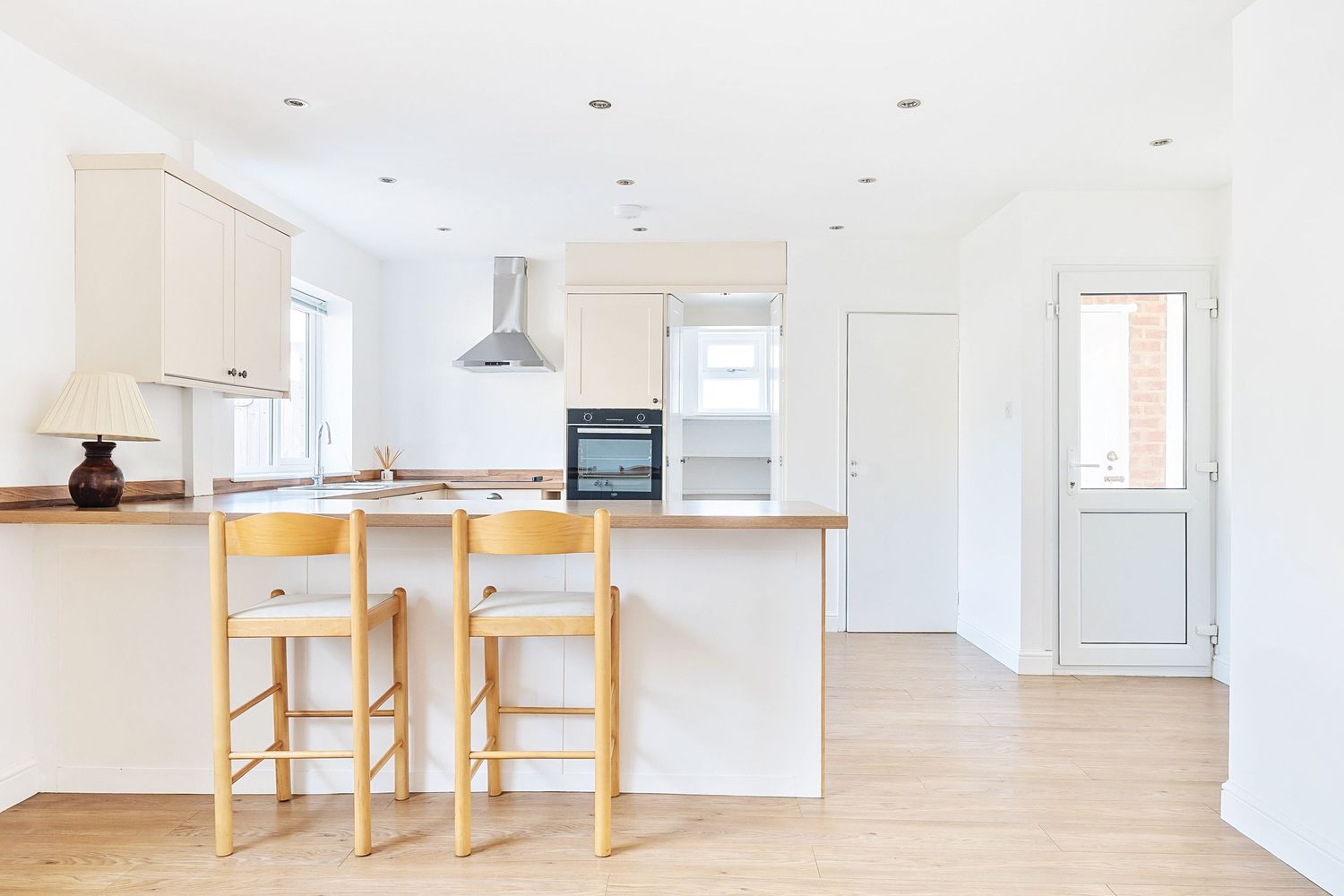 Modern white kitchen with wooden bar stools, butcher block countertop, and stainless steel hood vent in natural lighting.